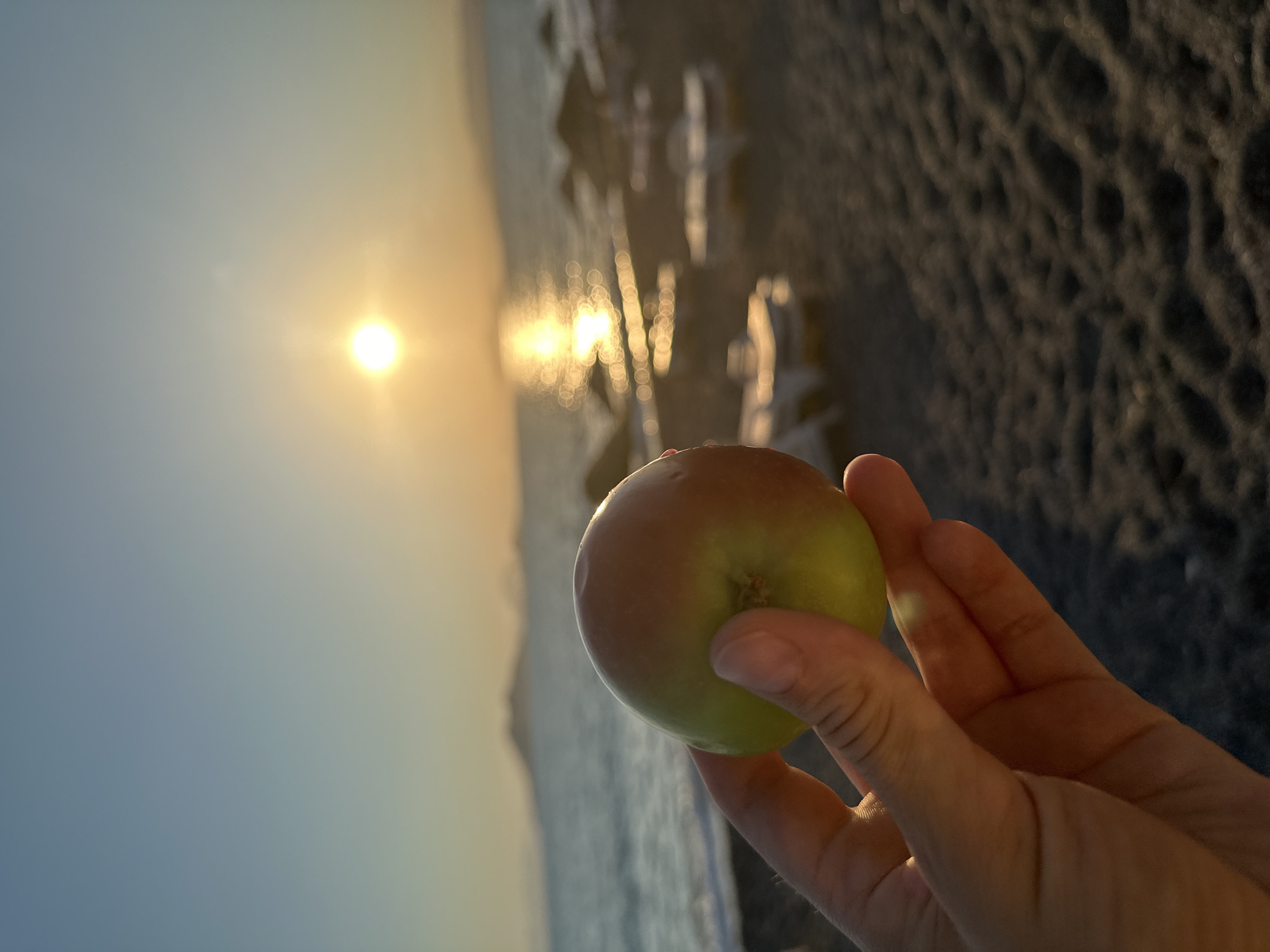 A person's hand holds a red and green apple in the foreground. The apple is slightly out of focus. In the background, a beach scene unfolds at sunset. The sun is low on the horizon, casting a golden glow on the water, which is choppy and reflects the sunlight in a shimmering path. The beach is made of dark pebbles or sand, with several empty lounge chairs and umbrellas visible. In the far distance, faint hills or islands can be seen. The sky is a gradient of pale blue to orange. The overall impression is a serene sunset at a beach location, likely in Fethiye, Turkey, given the context provided.
