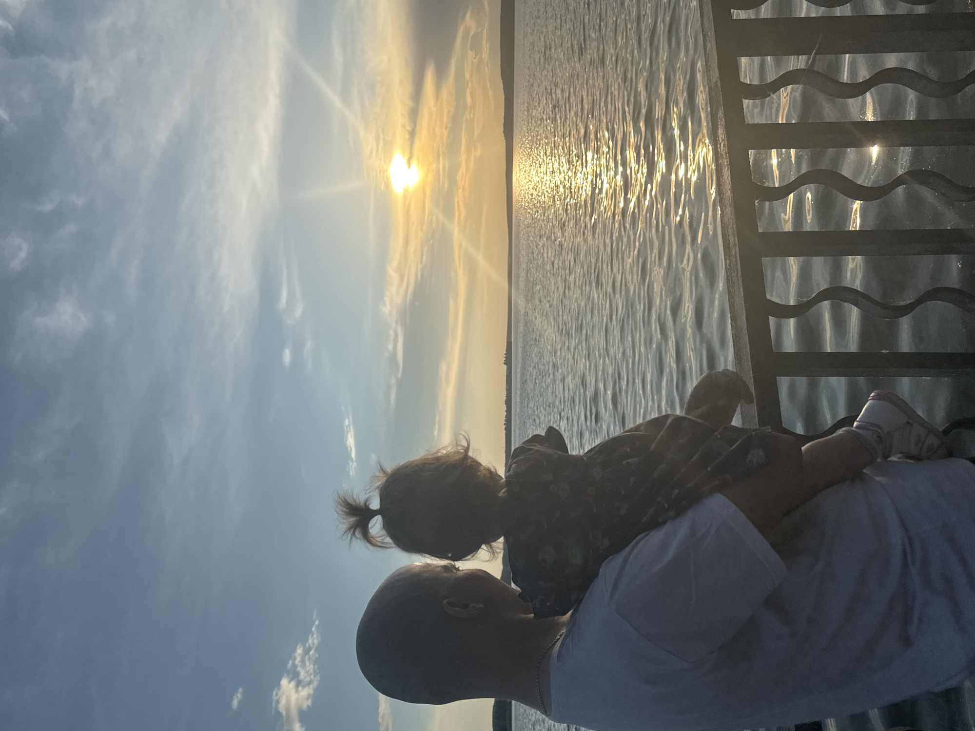 A person is holding a young child in their arms while facing a body of water at sunset. The sun is low in the sky, casting a warm glow on the water and clouds. The person appears to be standing on a railing overlooking the water. The child has their hair tied up in a small ponytail. The scene is likely a public area by the water, possibly a park or embankment, in the city of Votkinsk, Russia.