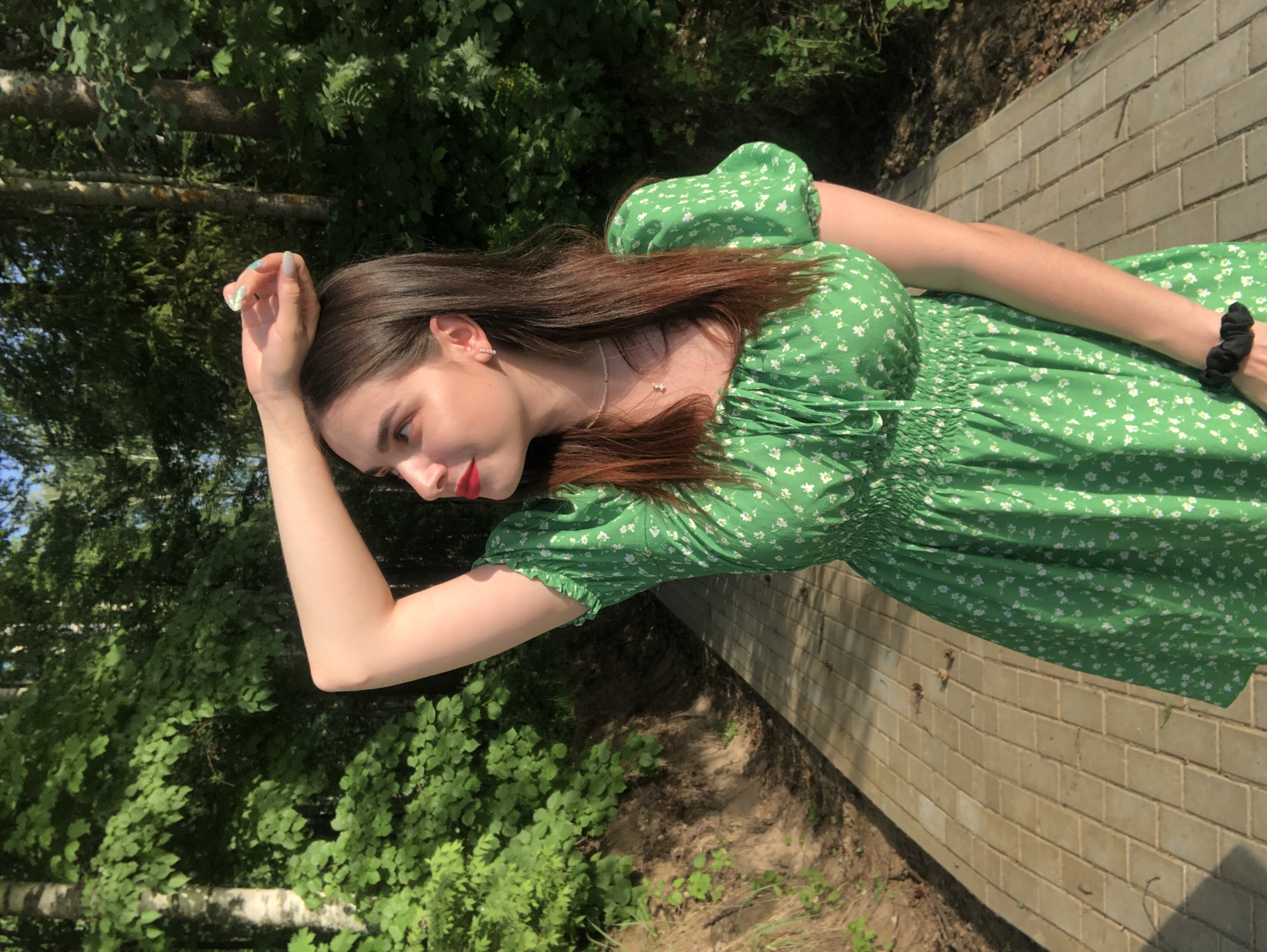 A young woman with long brown hair and a green floral dress is standing on a brick walkway outdoors. She is shielding her eyes with her left hand. The environment consists of trees and foliage. The location is Noviy, Russia.