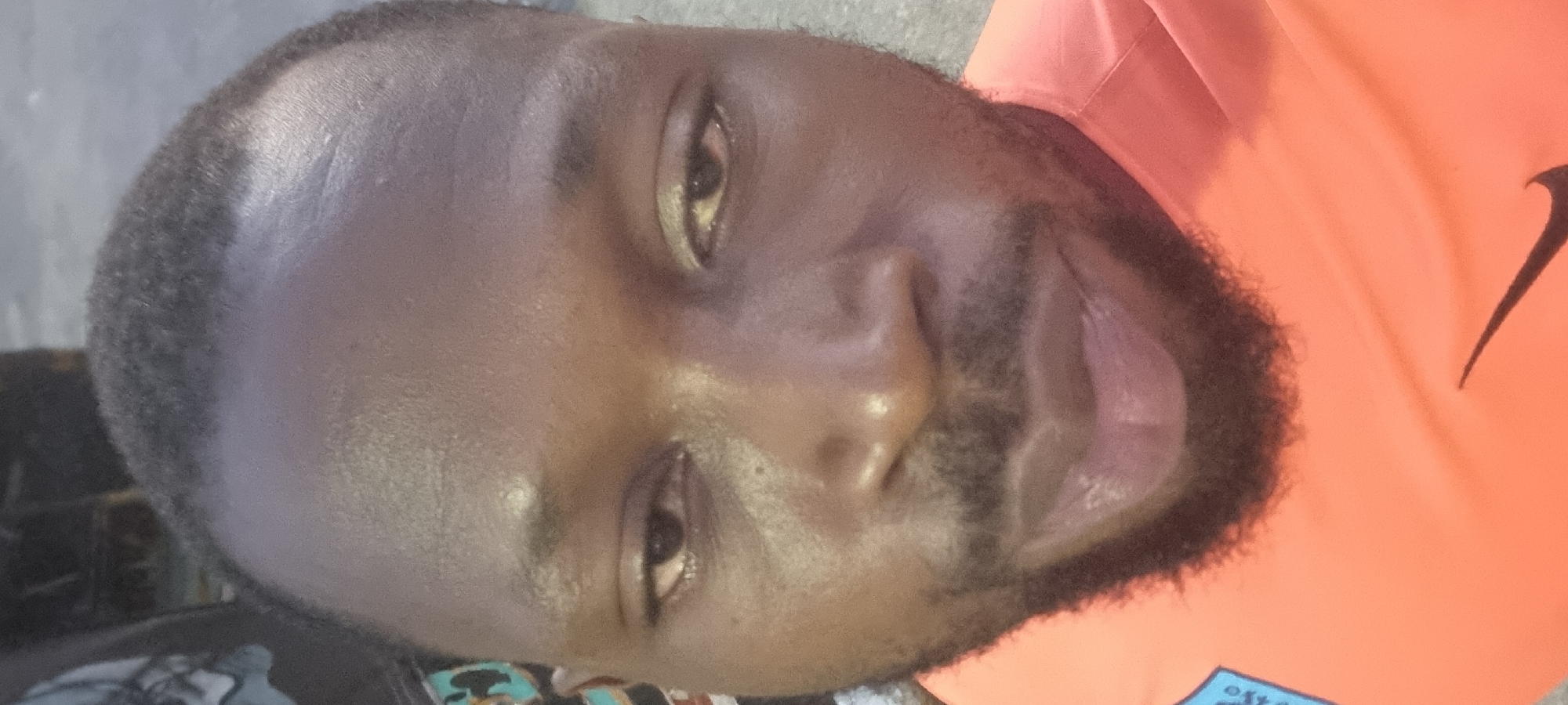 A close-up, rotated image shows a man with dark skin, short black hair, and a beard. He is wearing an orange shirt with a black Nike swoosh visible. The background is indistinct, but the setting is assumed to be Lagos, Nigeria.