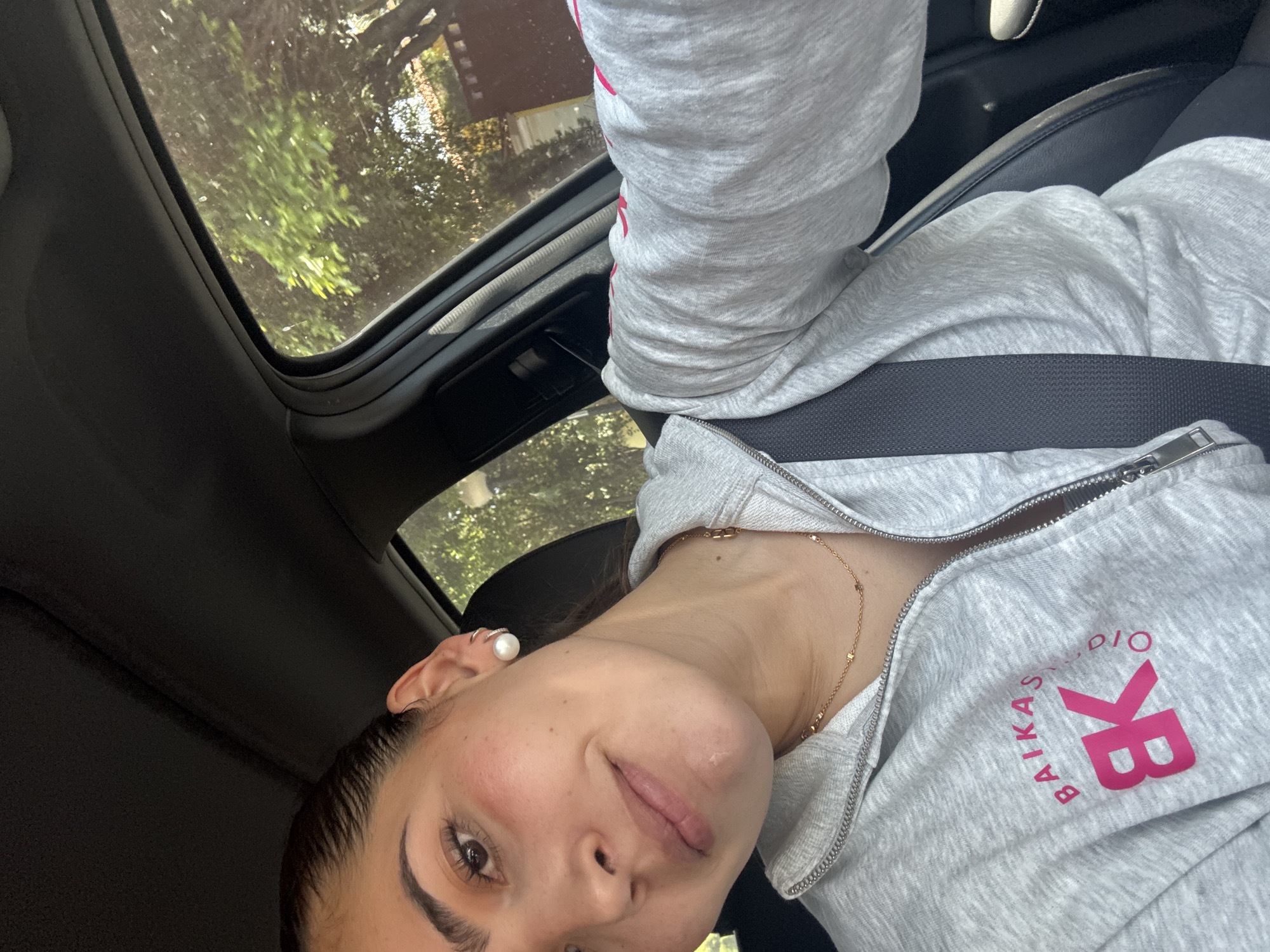 A selfie captures a young woman seated in a car, looking directly at the camera with a slight, playful smile, her tongue subtly visible. Her head is tilted, and she is wearing a light grey zippered hoodie. A prominent bright pink logo on the left chest of her hoodie reads "BAIKAS STUDIO" with a larger "BK" below it. A black seatbelt crosses her body, indicating she is secured in the vehicle. She accessorizes with a delicate gold chain necklace and a large pearl earring in her right ear.
The setting is inside a car with a dark interior. Above her, a sunroof reveals a view of green trees and foliage, suggesting it is daytime. The overall impression is a casual, candid moment taken while in transit, possibly within San Bernardino Tlaxcalancingo, Mexico, given the provided location context.