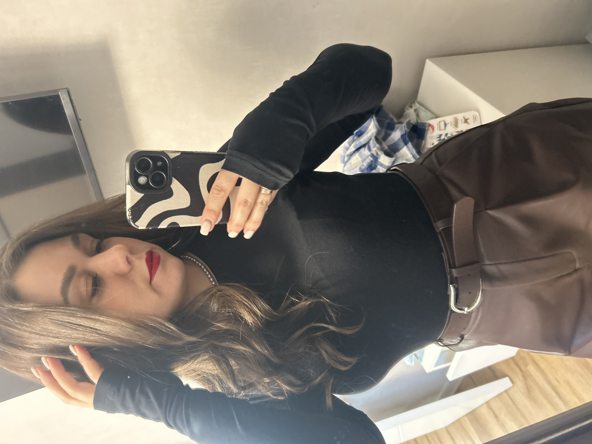 A woman with shoulder-length brown hair is taking a selfie indoors. She is wearing a black turtleneck top with extended sleeves that cover part of her hands. She has on brown leather pants with a brown belt. She is holding a phone with a black and white patterned case. The background includes a TV screen, a white shelf with items including a blue plaid shirt, and a wooden floor. The location is likely inside a home or apartment. It is not possible to determine the location in the city of Chaikovsky, Russia from the image alone.