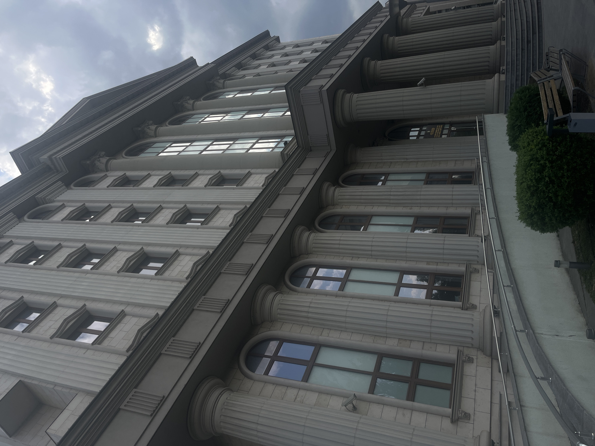 The image shows a large, neoclassical building with a grey facade and a cloudy sky above. The building features rows of tall, fluted columns and large, multi-paned windows. A ramp with metal railings leads up to the entrance, which is partially visible. In the foreground, there are green bushes and a wooden bench with a metal trash can. The scene is set in an urban environment, with no people visible. The lighting suggests it is daytime, though the overcast sky creates a muted atmosphere. The building's architecture indicates it is likely a public institution or office building.