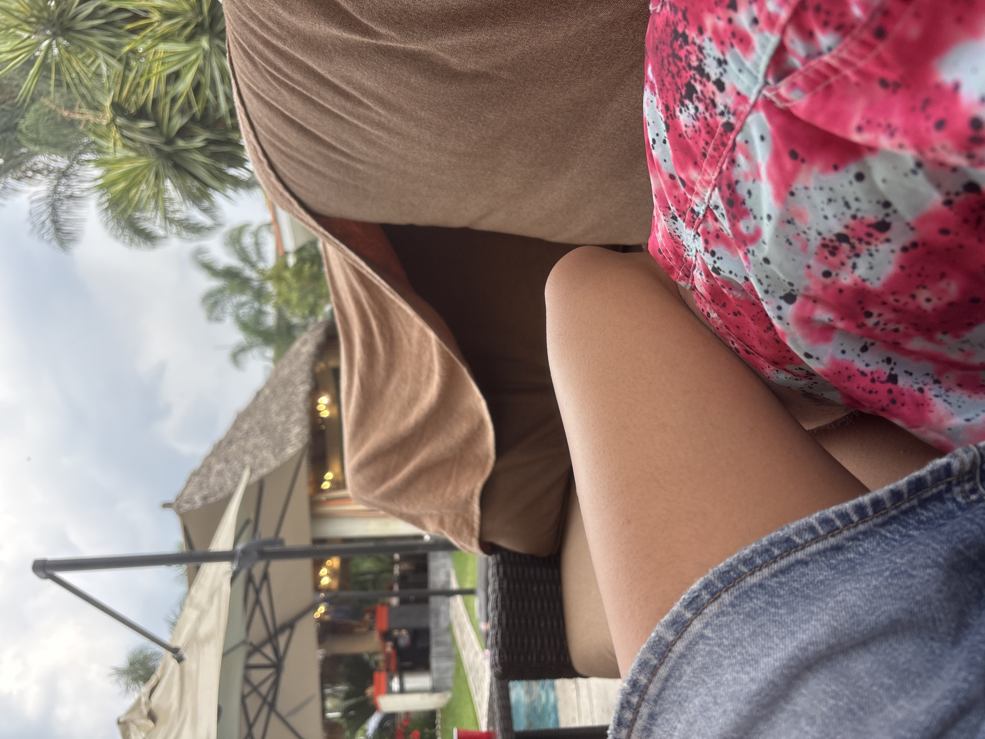 The image depicts a person seated outdoors, likely on a patio or veranda. The subject is wearing denim shorts and a pink and gray patterned hat. A portion of their leg is visible. They are seated on a brown cushioned outdoor sofa. In the background, there's a glimpse of an outdoor structure with a thatched roof, along with patio umbrellas and what appears to be a pool area. Palm trees are also visible, suggesting a tropical or subtropical location.