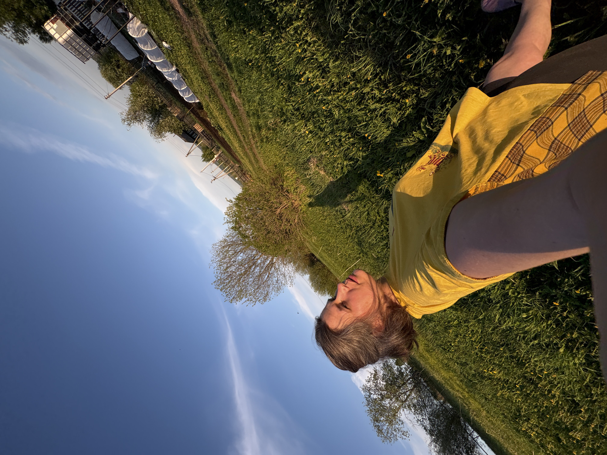 The image shows a person lying on a grassy field under a blue sky with wispy clouds. The person is wearing a yellow shirt and dark shorts. In the background, there are trees and what appears to be a structure covered with white material, possibly a greenhouse or agricultural setup. The photo was taken in the city of Nizhnyaya Chermoda, Russia. The perspective of the photo suggests it was taken from a low angle. The person is lying on their side.