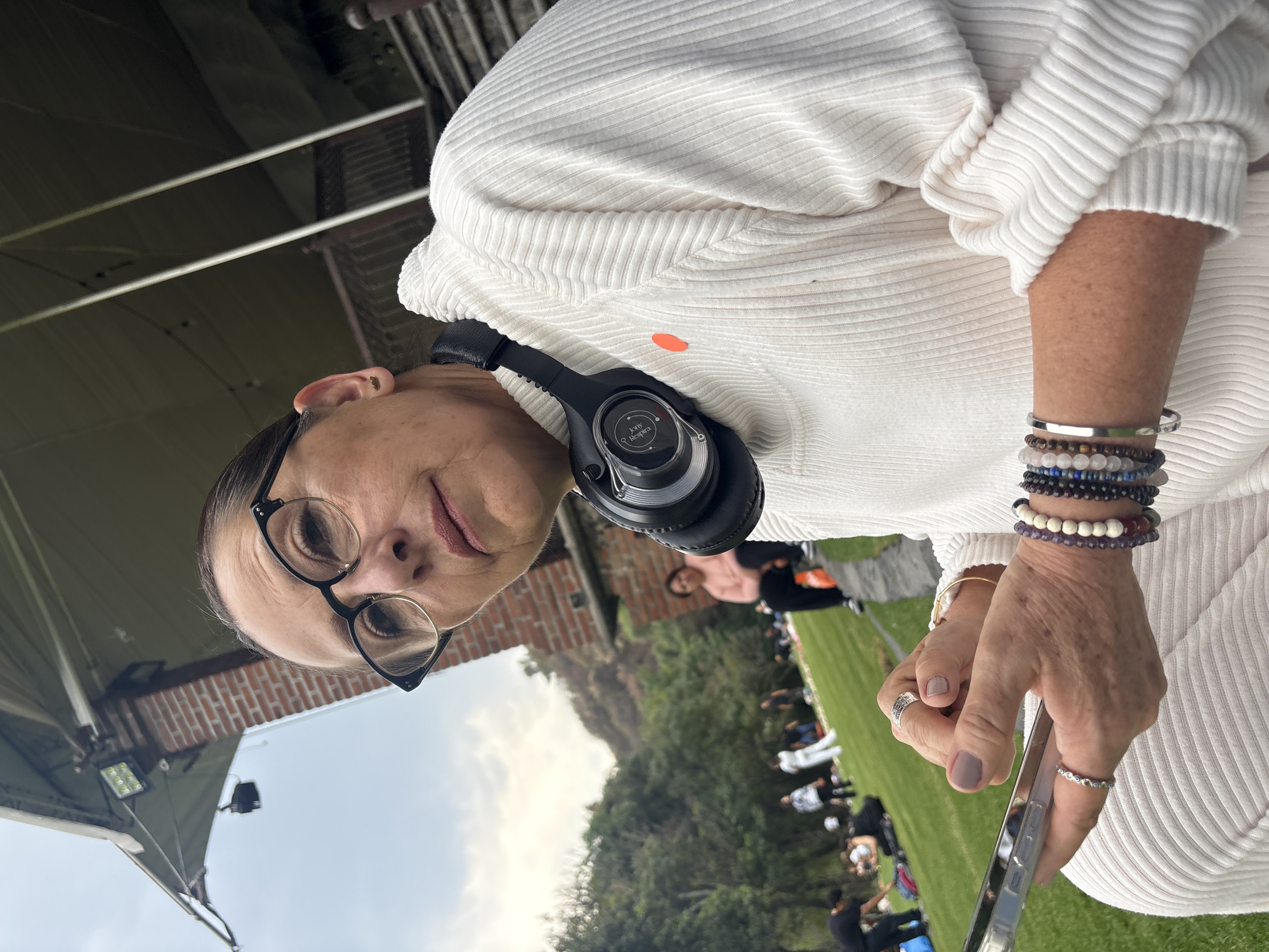 A woman in a white ribbed sweater and glasses is visible from the chest up. She has headphones around her neck and bracelets on her wrist. She is holding a smartphone. A grassy field with people sitting and standing is visible in the background. Trees and a mountain are also visible. The scene is located in Santa Ana Acozautla, Mexico.