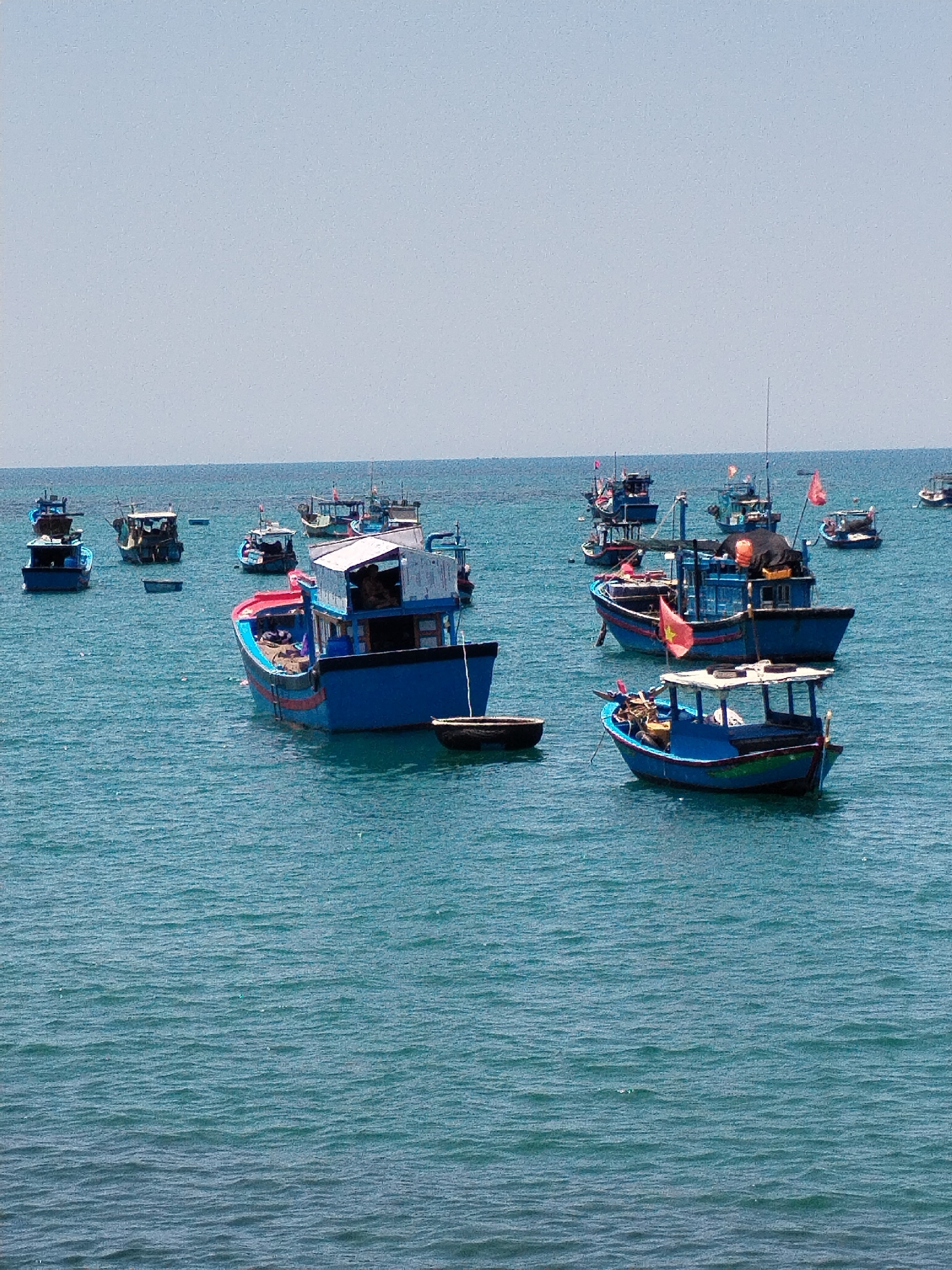 The image shows a harbor scene in Bac Nha Trang Ward, Vietnam, with several blue fishing boats floating on the water. Some of the boats have masts and flags. The sky is pale and clear. The sea stretches out to the horizon, and the boats are scattered across the water.