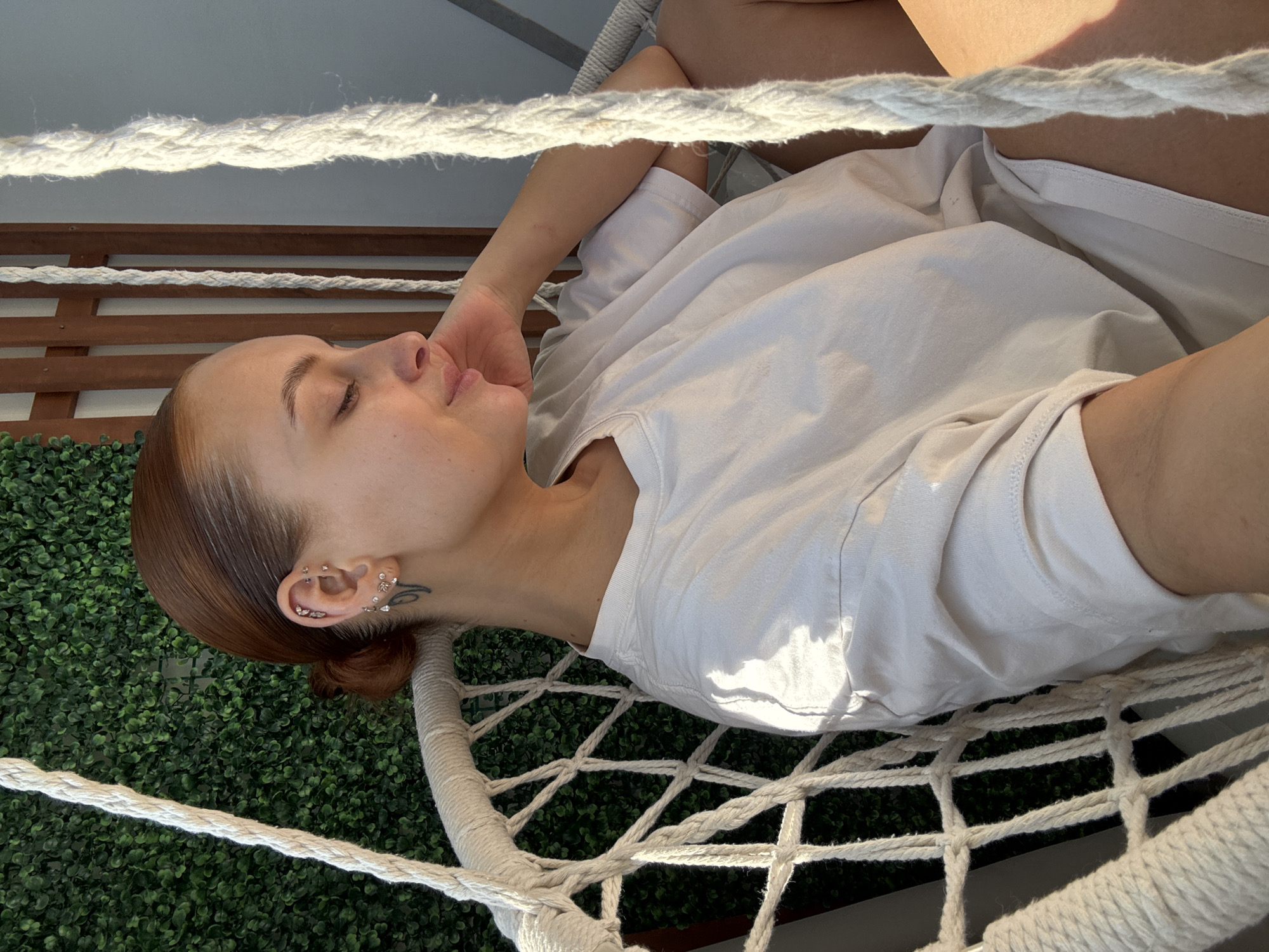 A young woman is reclining in a macrame hammock chair on a balcony. She is fair-skinned with auburn hair pulled back in a low bun, wearing a white t-shirt, and resting her head on her hand. Her eyes are closed. The background behind the hammock chair consists of a faux ivy wall. The setting may be located in the city of Chaikovsky, Russia.