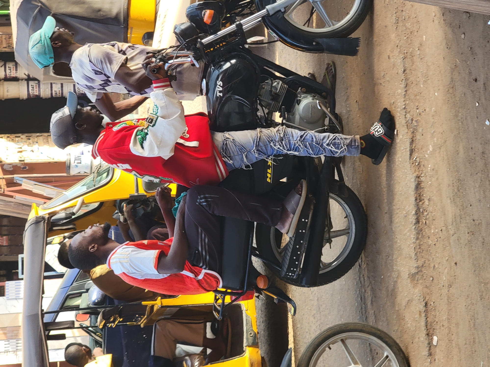 A medium-angle shot shows three men on a motorcycle and in a yellow vehicle in what appears to be an urban setting. One man, wearing a red jacket and white cap, sits on the back of the motorcycle, his feet visible in open-toed sandals. Another man, wearing a patterned shirt and a blue cap, is positioned on the motorcycle towards the front. The motorcycle is black with a silver engine, and its license plate reads "KLZ". A third man sits in a yellow vehicle with a red and white shirt, partially visible. Additional people are seated inside the yellow vehicle. The background reveals other vehicles and buildings, suggesting a busy street in Maiburiji, Nigeria. The road surface is visible.