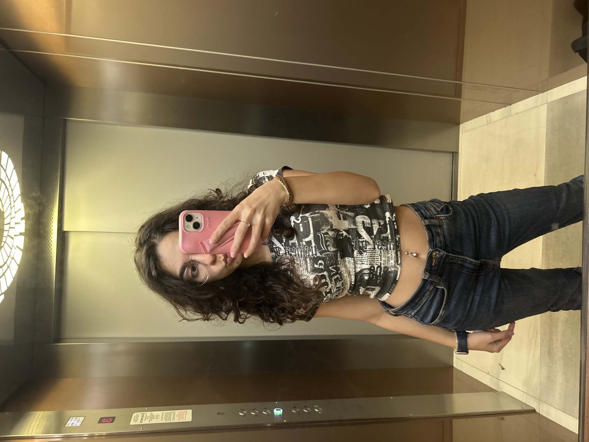 A woman is taking a selfie in an elevator. She is wearing jeans, a cropped t-shirt with black and white graphics, and a watch. She has curly hair and wears glasses. The phone she is using is pink. The elevator has a metal interior with a row of buttons and a light fixture on the ceiling. It is unknown if this is in the city of Sofia, Bulgaria.