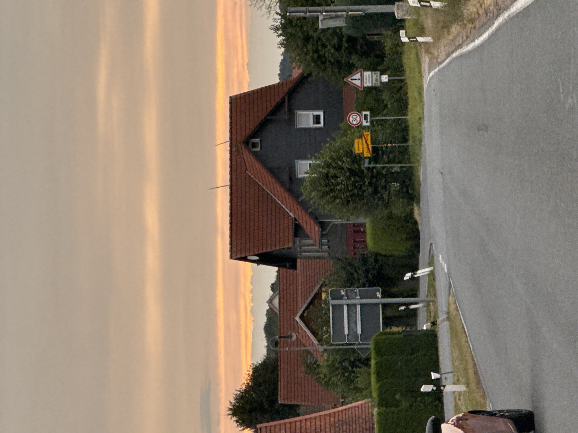 A paved road curves through a village in Hohenstein, Germany, during sunset. The sky displays streaks of orange and pink. On the left side of the road, a car is partially visible. The right side features a manicured hedge, road markers, and several traffic signs, including a speed limit sign indicating 30. Beyond the hedge, houses with red tile roofs and dark-colored facades are nestled among trees.