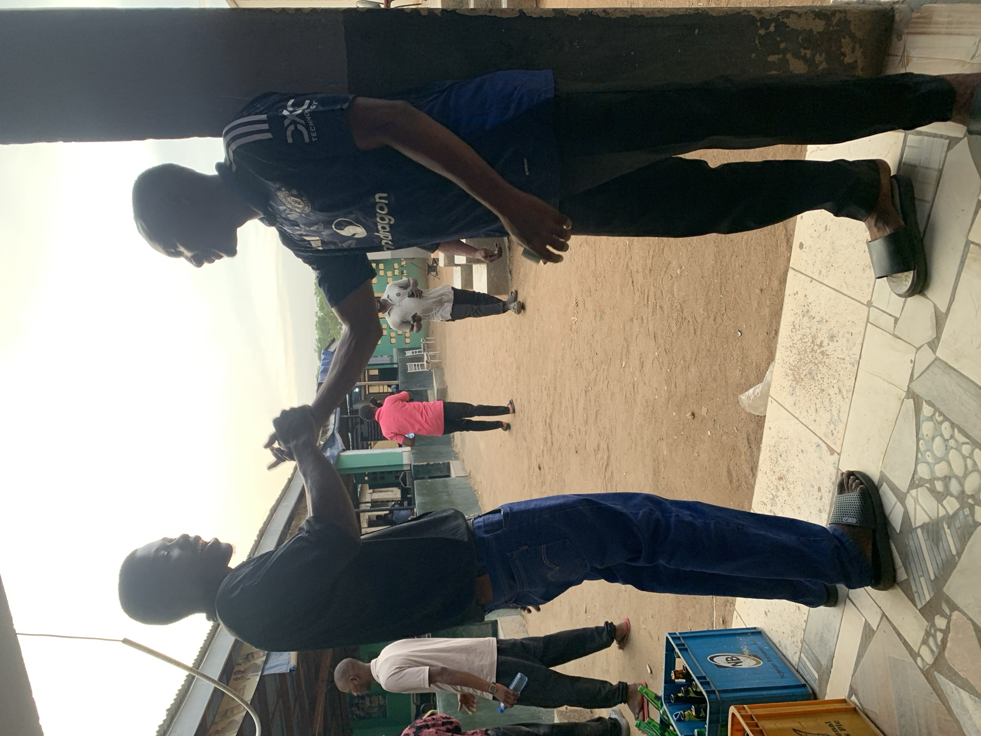 Two men stand beneath a corrugated metal awning, their hands clasped together in a handshake or celebratory gesture. The man on the left wears a black shirt and dark blue jeans, while the man on the right sports a dark blue jersey with white stripes and black pants. Both men wear sandals.
In the background, several people are visible on a dirt ground. One person wears a pink shirt. Another individual is wearing a white shirt and holding a blue water bottle.
A blue crate filled with green bottles and another yellow/orange crate are near the man on the left. The ground beneath the awning is tiled with a patterned design. The location is identified as Kabong, Nigeria.