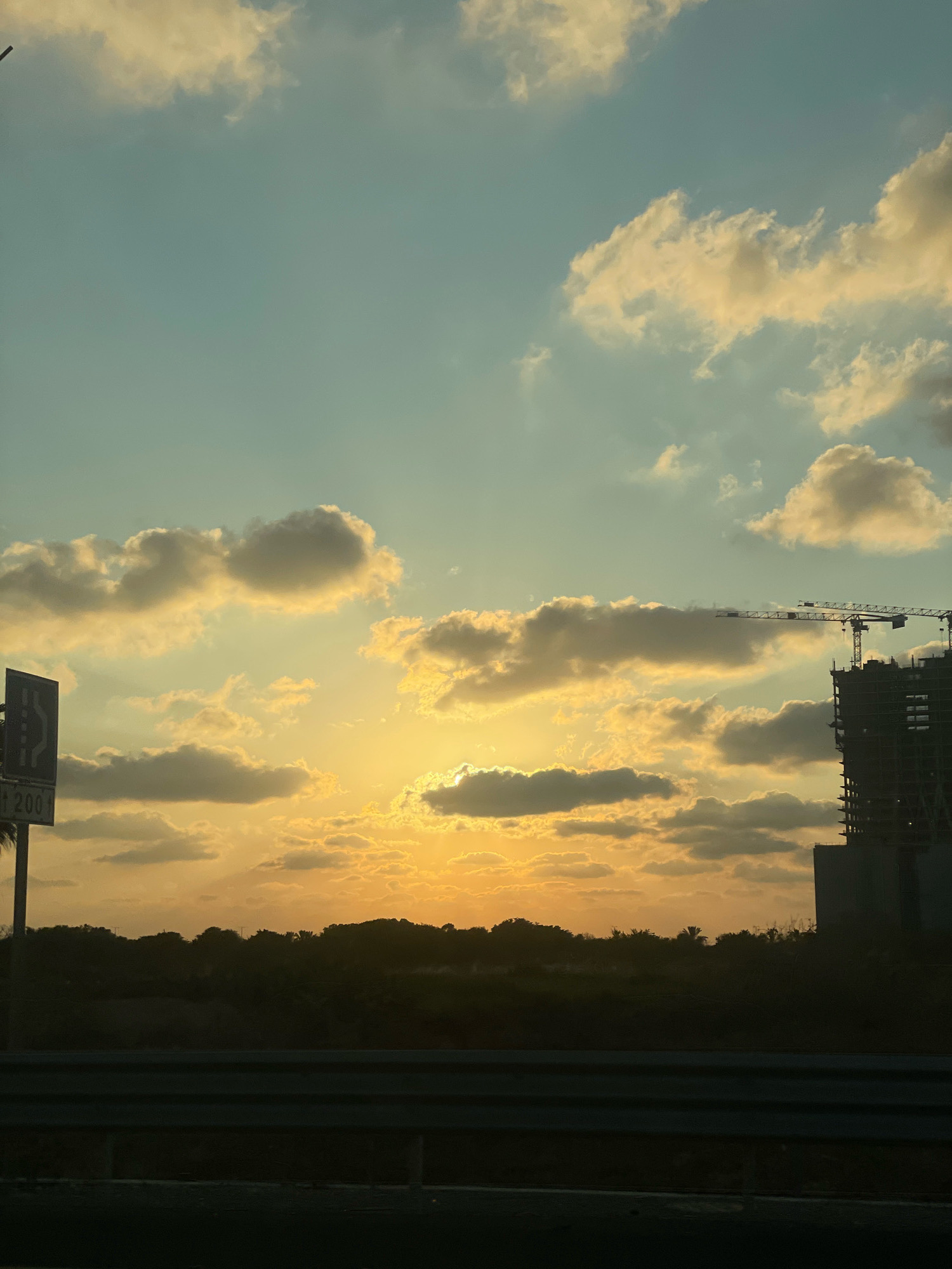 The image depicts a sunset over a construction site in Herzliya, Israel. The sky is filled with a mix of pale blue and golden-yellow clouds, illuminated by the setting sun which is partially obscured by clouds. In the distance, silhouetted against the bright sky, is a developing building with construction cranes positioned on top, indicating ongoing development. The foreground shows a dark landscape, possibly with trees and vegetation, and a metal barrier or railing. A road sign with Arabic and Hebrew text is visible on the left side of the frame. The overall scene suggests an urban or suburban environment experiencing rapid growth, captured during the tranquil beauty of sunset.