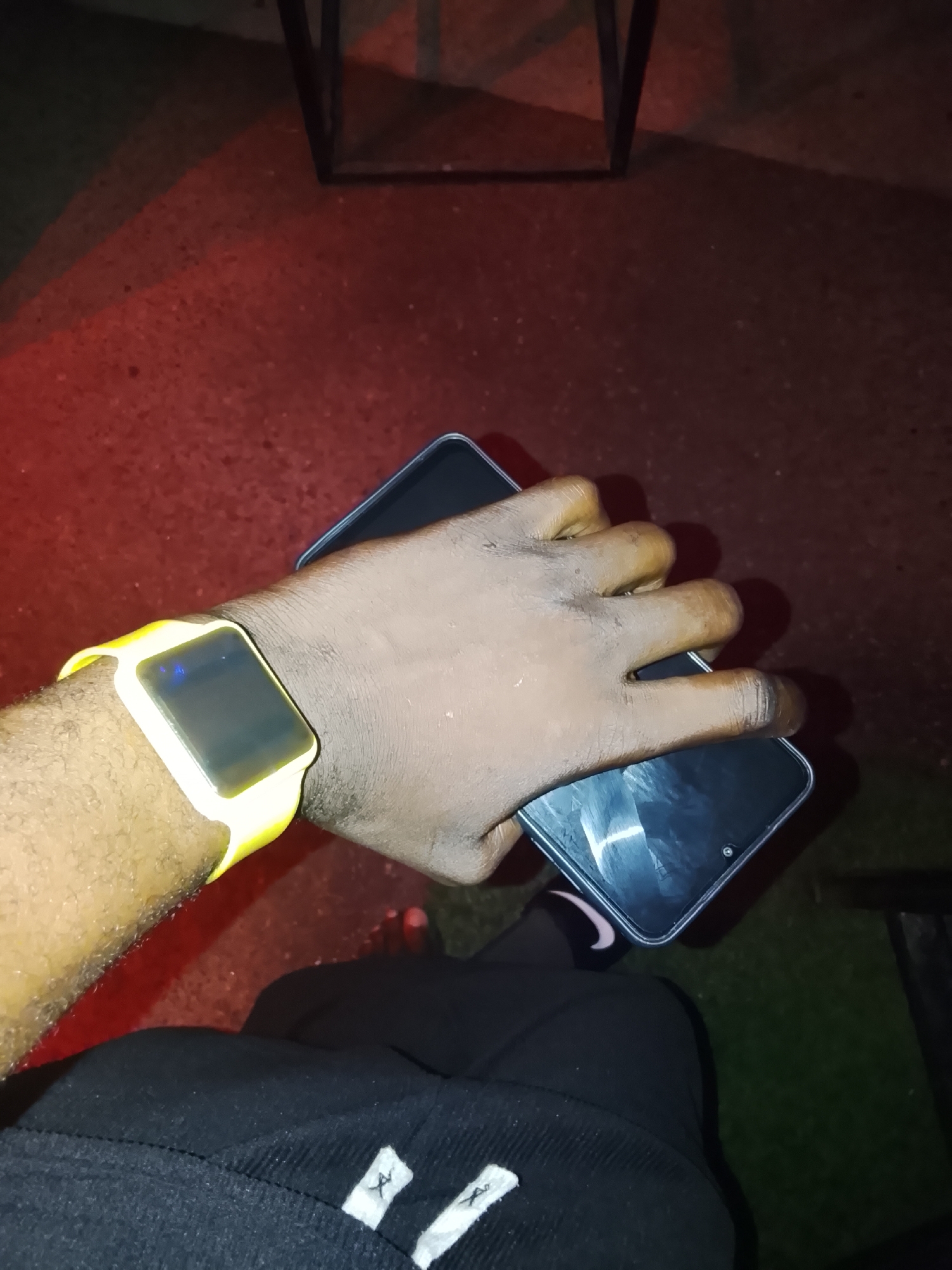 A close-up, high-angle shot shows a person's left arm and hand wearing a bright yellow smartwatch and holding a black smartphone. The skin tone is dark. The smartwatch has a dark screen with a blue light reflecting on it. The phone is held horizontally, with the person's fingers wrapping around its side. The background is dark and appears to be an outdoor setting with a red-toned ground and some green grass. There are shadows visible, suggesting artificial lighting. No specific landmarks or details confirm the location as Howayi, Nigeria.
