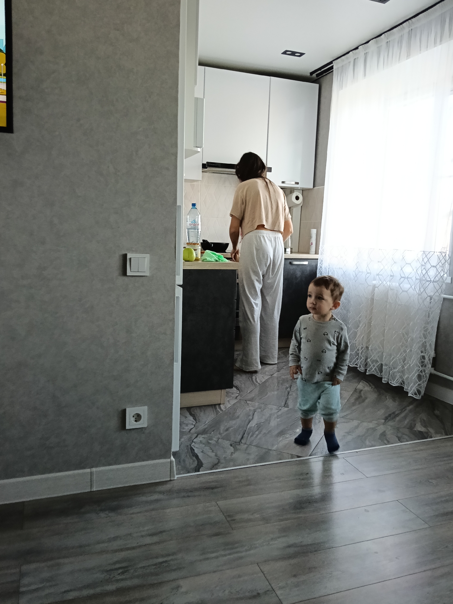 A child stands in the foreground of a domestic scene in Murom, Russia. The child wears a grey long-sleeved shirt with a pattern and light blue shorts, and dark blue socks. Behind the child, a woman with short dark hair is at a kitchen counter, possibly cooking. The kitchen has white cabinets and a dark countertop, and a bottle of "Опдаг" water is visible on the counter. A white curtain covers a window to the right. The flooring transitions from a grey-toned wood in the foreground to a grey marble pattern in the kitchen area. A grey wall is visible in the left side of the frame, featuring light switches and an outlet.