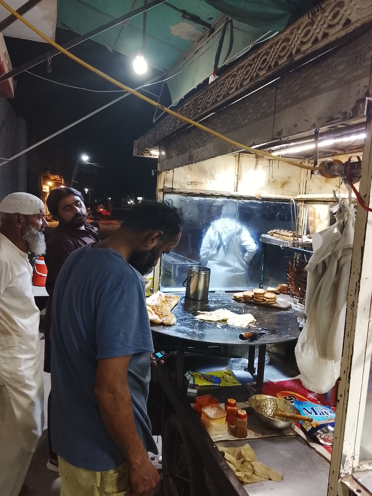 This vertical-format image captures a vibrant street food stall scene at night. Three men are gathered around a bustling cooking area, where food is being prepared on a large, circular griddle (tawa).
In the foreground and midground, three men observe the cooking process. On the far left, an older man with a white skullcap (kufi) and a long white beard wears a light-colored outfit. Next to him, a man with a dark beard and hair, dressed in a dark shirt, looks intently at the food. The third man, central in the image, also has a dark beard and is wearing a blue t-shirt and light-colored shorts or trousers; he is leaning forward, focused on the griddle.
Behind the griddle, inside the stall, another person in a white garment is visible, appearing to be the cook or an assistant. On the griddle, various items are cooking, including flatbreads or parathas, some appearing to be filled, and a stack of round patties or burgers. A metal pot and spatula are also on the cooking surface. Below the main counter, a shelf holds condiments such as red and yellow squeeze bottles (likely sauces like ketchup and chili) and spice shakers. A packet clearly labeled "May" (likely Mayonnaise) is also visible.
The stall itself is brightly lit by bare hanging light bulbs, contrasting with the dark night sky. A green awning covers the top, and a yellow pipe runs across the upper part of the frame. Decorative metalwork adorns the upper beam of the stall's structure. Streetlights are faintly visible in the dark background, further confirming the nighttime setting. The men appear engaged and observant, suggesting they are customers waiting for or watching their food being made.