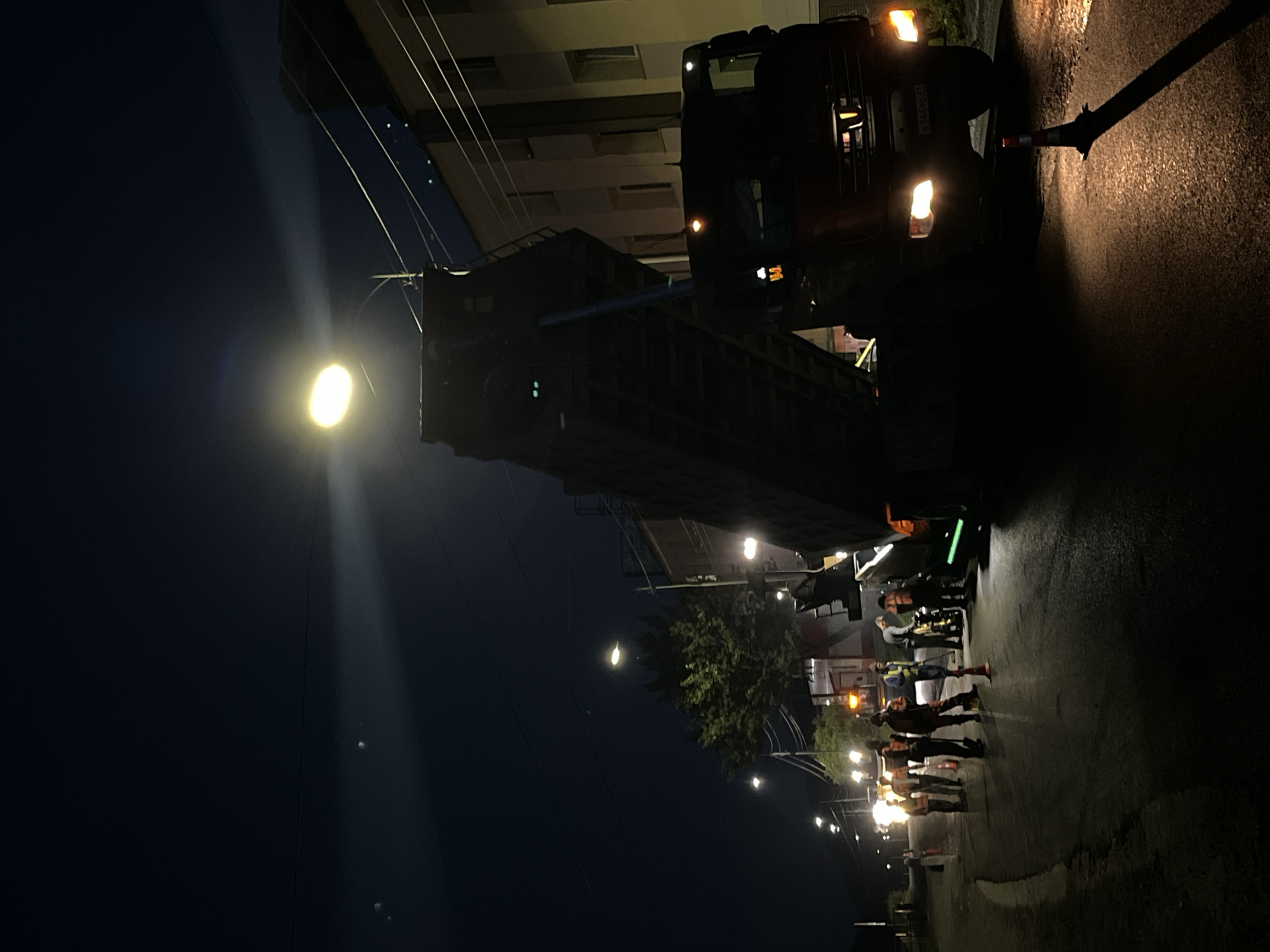 At night, on a street in Petropavlovsk-Kamchatsky, Russia, a group of road workers are engaged in what appears to be road repair. A large dump truck, with its bed raised, is positioned on the road, likely to offload material. Several individuals, wearing reflective vests, are standing near the truck, some holding tools. Streetlights and the truck's headlights illuminate the scene. The surrounding environment suggests a residential or commercial area, with buildings visible in the background and trees lining the street. The road surface appears wet, possibly from recent rain or cleaning. The overall atmosphere is one of active construction work occurring after dark.
