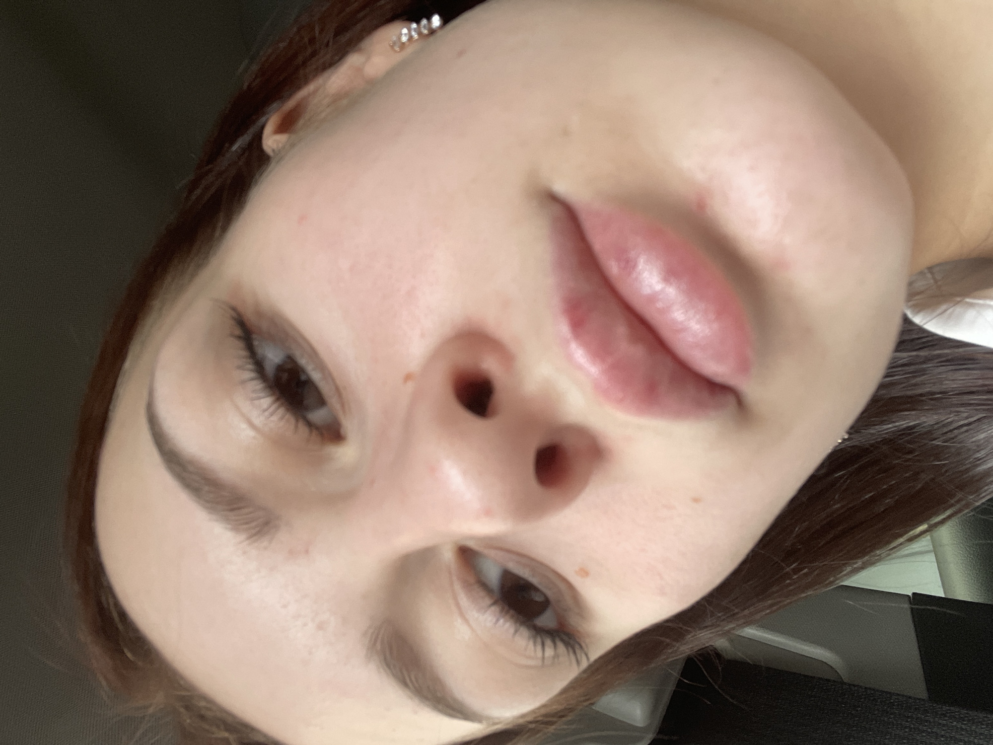 The image is a close-up, high-angle, indoor, eye-level shot of a young Caucasian female's face. Her head is tilted downwards and to her left. She has auburn hair, prominent eyebrows, long eyelashes, and light brown eyes. Her lips are full and appear to be glossed. There are a few freckles visible on her cheeks and nose, as well as some redness and small blemishes on her skin. She is wearing a small, silver earring with multiple stones in her right ear. The background is blurred and dark, suggesting an indoor environment with low lighting. The image quality is moderately good, with some graininess. There is no discernible indication of the city of Semyonov, Russia.