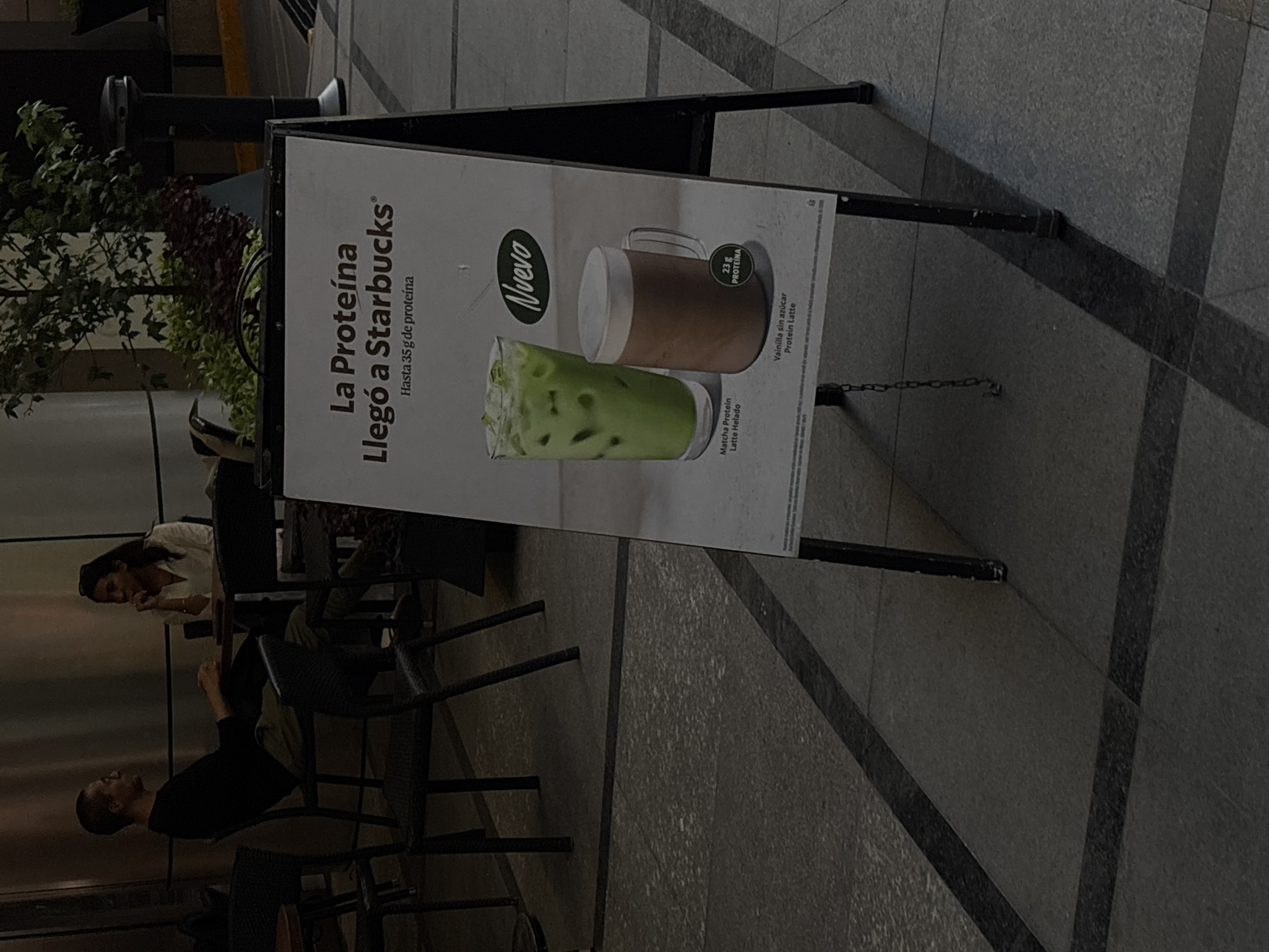 The image shows a promotional sign for Starbucks in Mexico City, Mexico. The sign is an A-frame board displaying an advertisement in Spanish, "La Proteína Llegó a Starbucks" (Protein Arrived at Starbucks), highlighting the protein content of their drinks (up to 35g). The ad features images of a "Matcha Protein Latte Helado" (Matcha Protein Iced Latte) and a "Vainilla sin azúcar Protein Latte" (Sugar-free Vanilla Protein Latte), noting the latter contains 23g of protein. In the background, two people are seated at outdoor cafe furniture near the signage. The location appears to be a public area with tiled flooring.