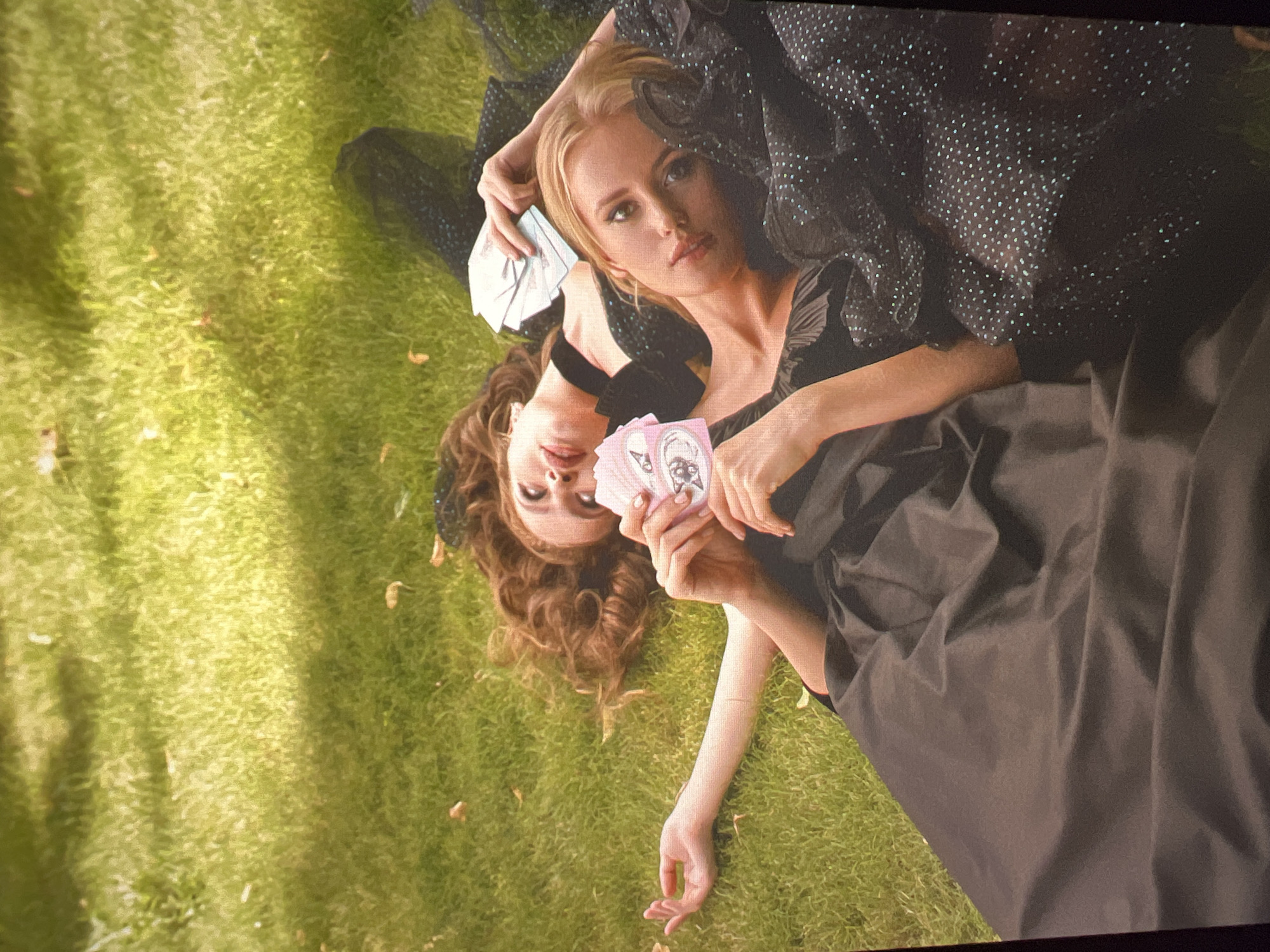 Two women in black dresses are lying on a grassy lawn in Saint Petersburg, Russia. The woman in the foreground has blonde hair and is looking at the camera. The woman behind her has red hair and is looking away from the camera. Both women are holding playing cards. The woman in the foreground is wearing a black dress with a sparkly overlay, and the woman in the background is wearing a simpler black dress. The lawn is green and appears to be in a park or garden. There are some fallen leaves scattered on the grass. The lighting suggests it is daytime, possibly with dappled sunlight.