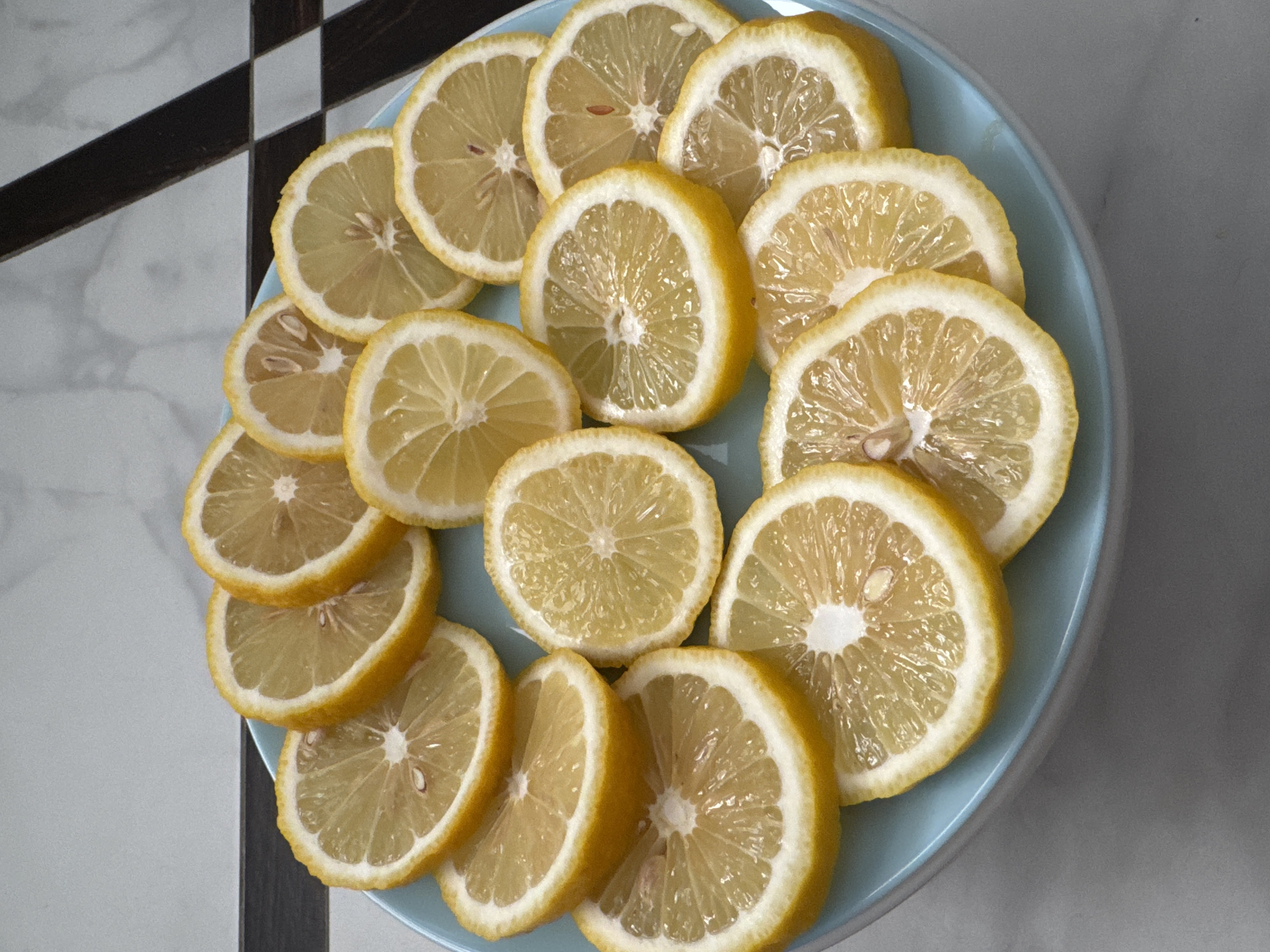 A light blue plate holds numerous slices of lemons. The lemon slices are arranged in a fanned pattern, with some overlapping. The background is a tiled surface with a pattern of white and dark grey squares. The image is taken from a top-down perspective. There are no people or animals present in the image. The scene is static and appears to be a still life photograph of food. The location is not specified in the image, so it cannot be confirmed as Mikhaylovsk, Russia.