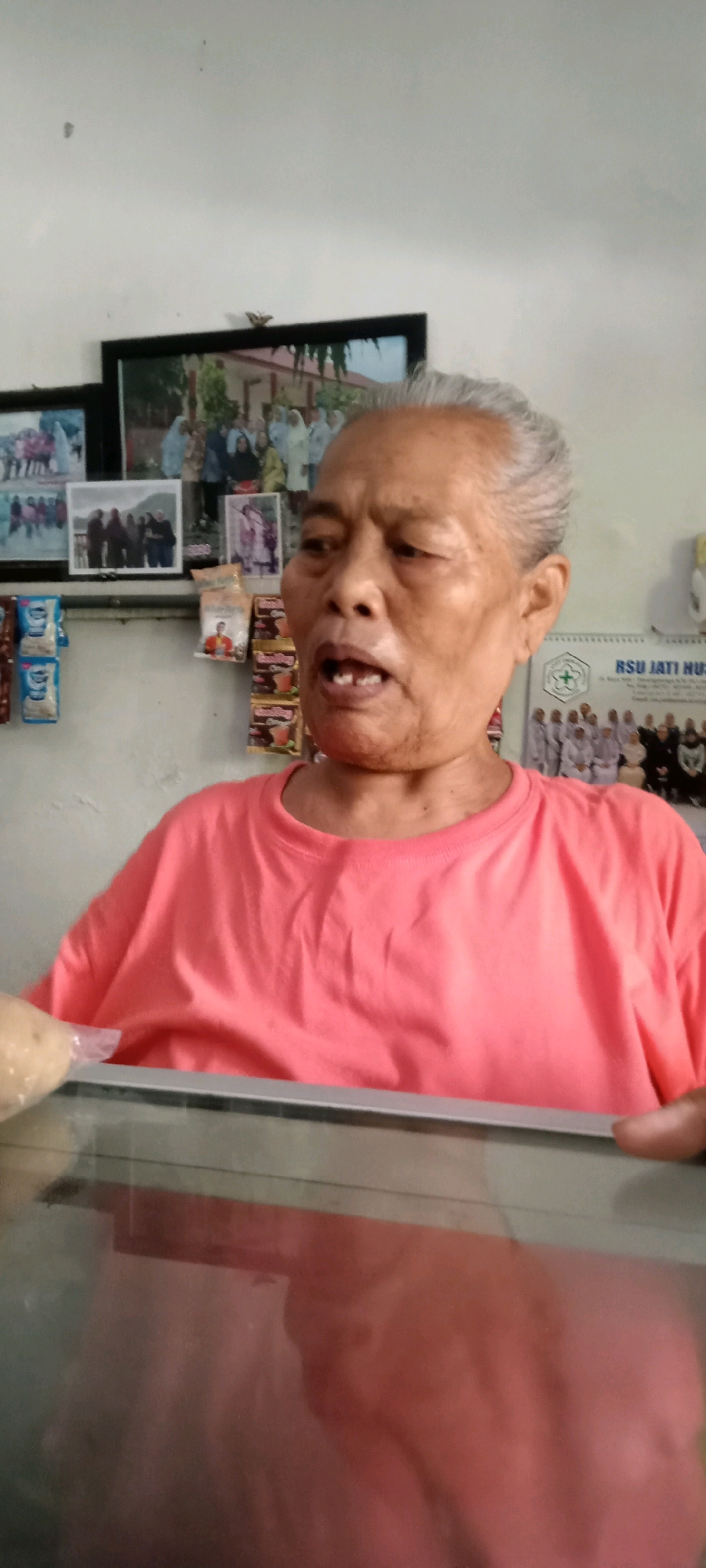 The image shows a close-up of an elderly woman with grey hair, wearing a bright pink t-shirt. She is looking to her right with her mouth open, revealing her teeth. Behind her, on a white wall, are several framed photographs. To the right of the photographs, a calendar with the text "RSU JATI NU" is visible. The scene appears to be indoors, possibly in a home or a small shop, given the display of packaged goods on a shelf below the photographs. A clear, reflective surface, likely a glass counter or display case, is in the foreground, partially obscuring the woman's lower body and shirt. The background elements suggest the location is in Jaten, Indonesia, as indicated by the text on the calendar.