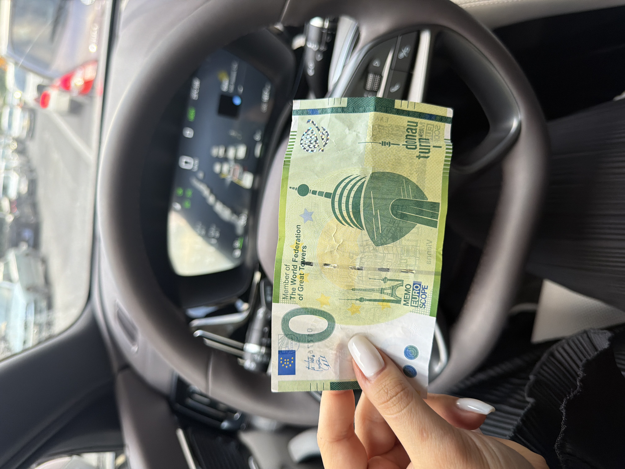 The image shows a close-up of a "0 Euro" souvenir banknote featuring the Donauturm (Danube Tower) in Vienna, Austria, being held in front of the steering wheel of a vehicle. The hand holding the note has a pale skin tone and long, white, manicured nails. The vehicle's dashboard and steering wheel are visible in the background, with the car's digital display showing blurred information. Outside the vehicle's windshield, there is a blurry view of a road and other cars, suggesting the vehicle is parked or stationary on a street. The background environment is not clear enough to identify the location as Yerevan, Armenia.