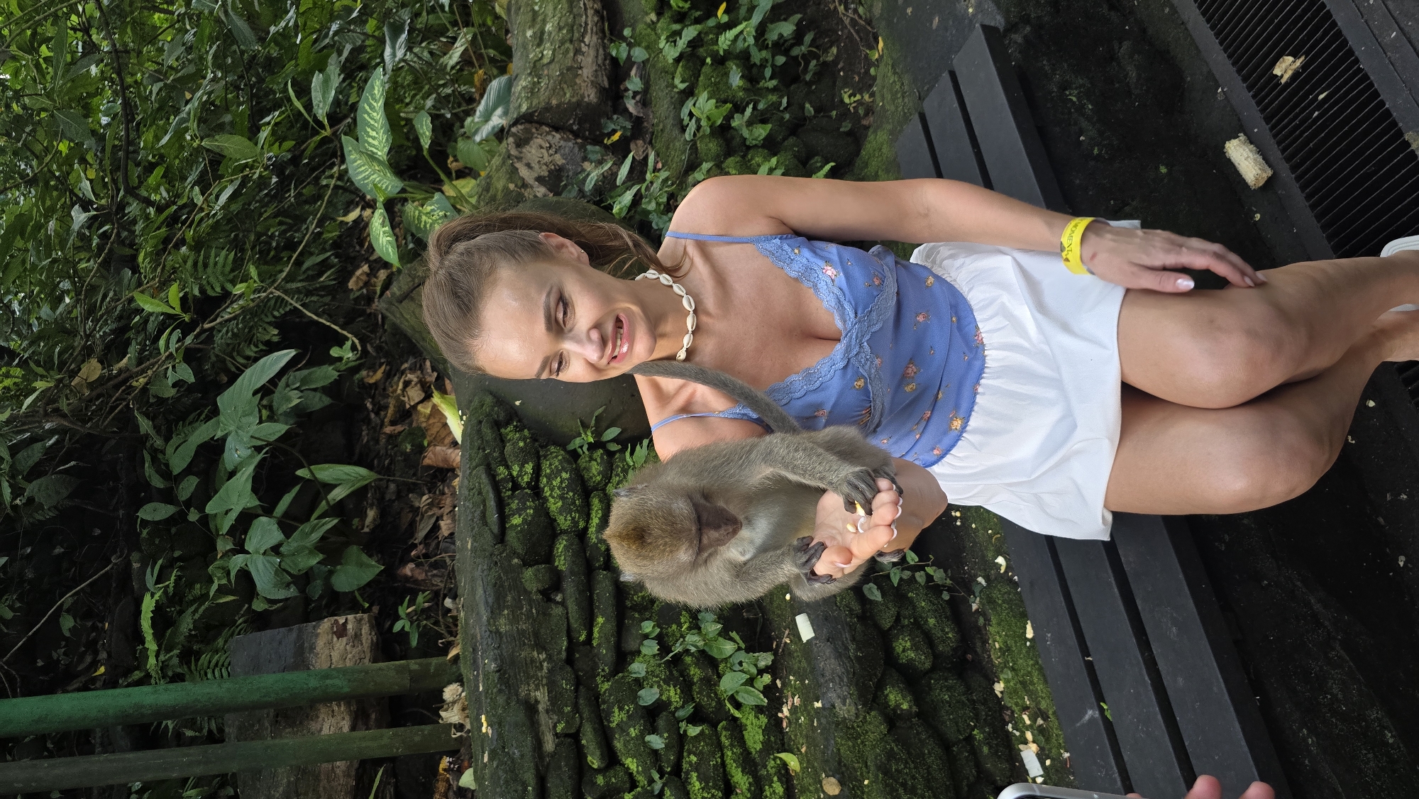 The photo captures a scene in the Ubud District of Indonesia, likely the Sacred Monkey Forest Sanctuary. A woman with brown hair pulled back in a ponytail sits on a wooden bench, wearing a blue top with lace trim, a white skirt, and a shell necklace. She is smiling and looking down at a monkey that is holding her foot. The environment is lush with tropical vegetation, including moss-covered rocks and various plants. A yellow wristband is visible on her wrist, and a portion of a cell phone held by a person off-camera is seen at the bottom of the frame.