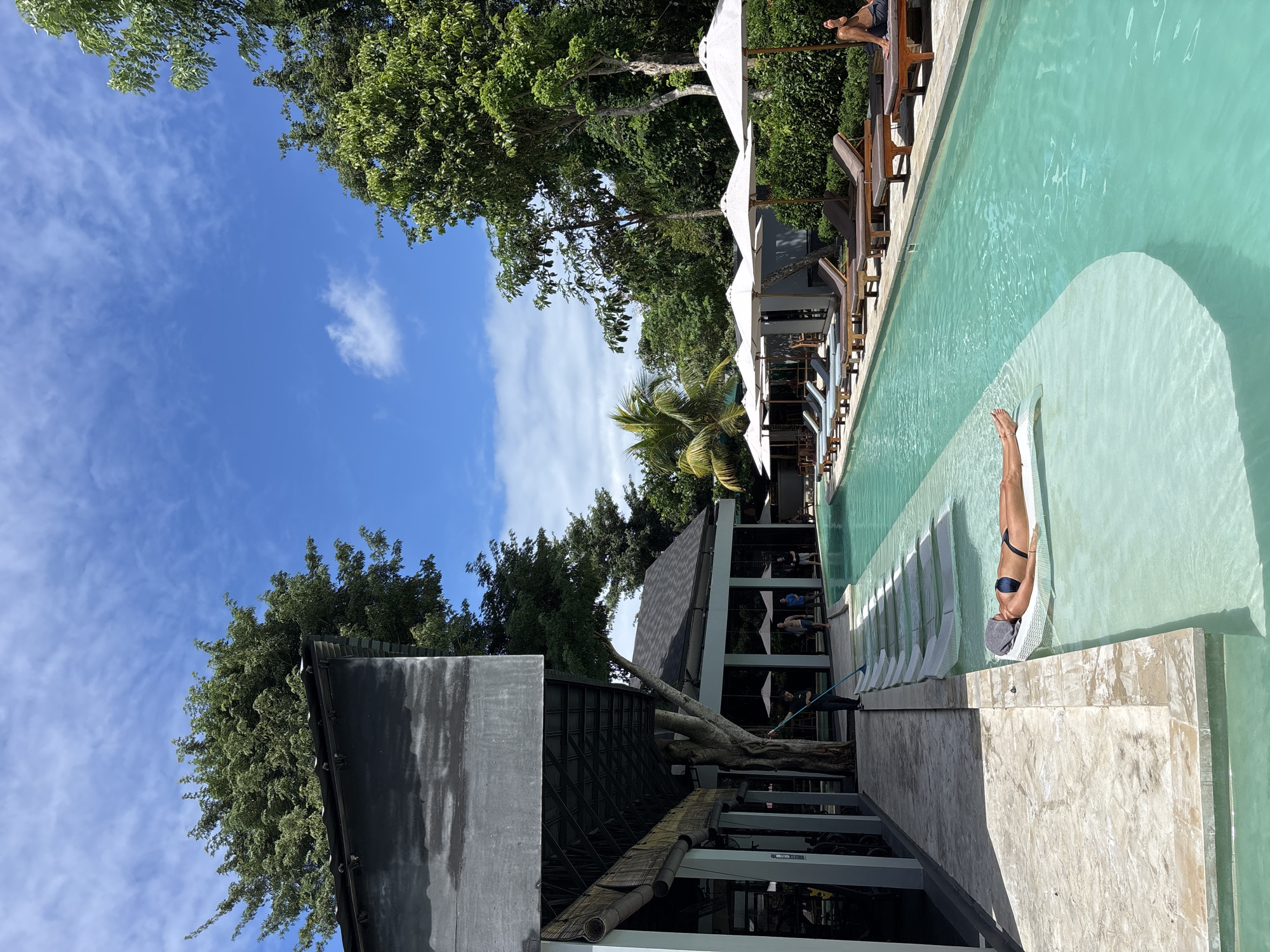 The image shows an outdoor swimming pool at a resort in Kuta Selatan, Indonesia. The pool is a light green color with white steps leading into the water. A woman in a black swimsuit and grey swim cap is lying in the water on the steps. There are lounge chairs with white umbrellas next to the pool. The resort building is to the left of the pool. The sky is blue with some white clouds, and there are trees surrounding the resort.