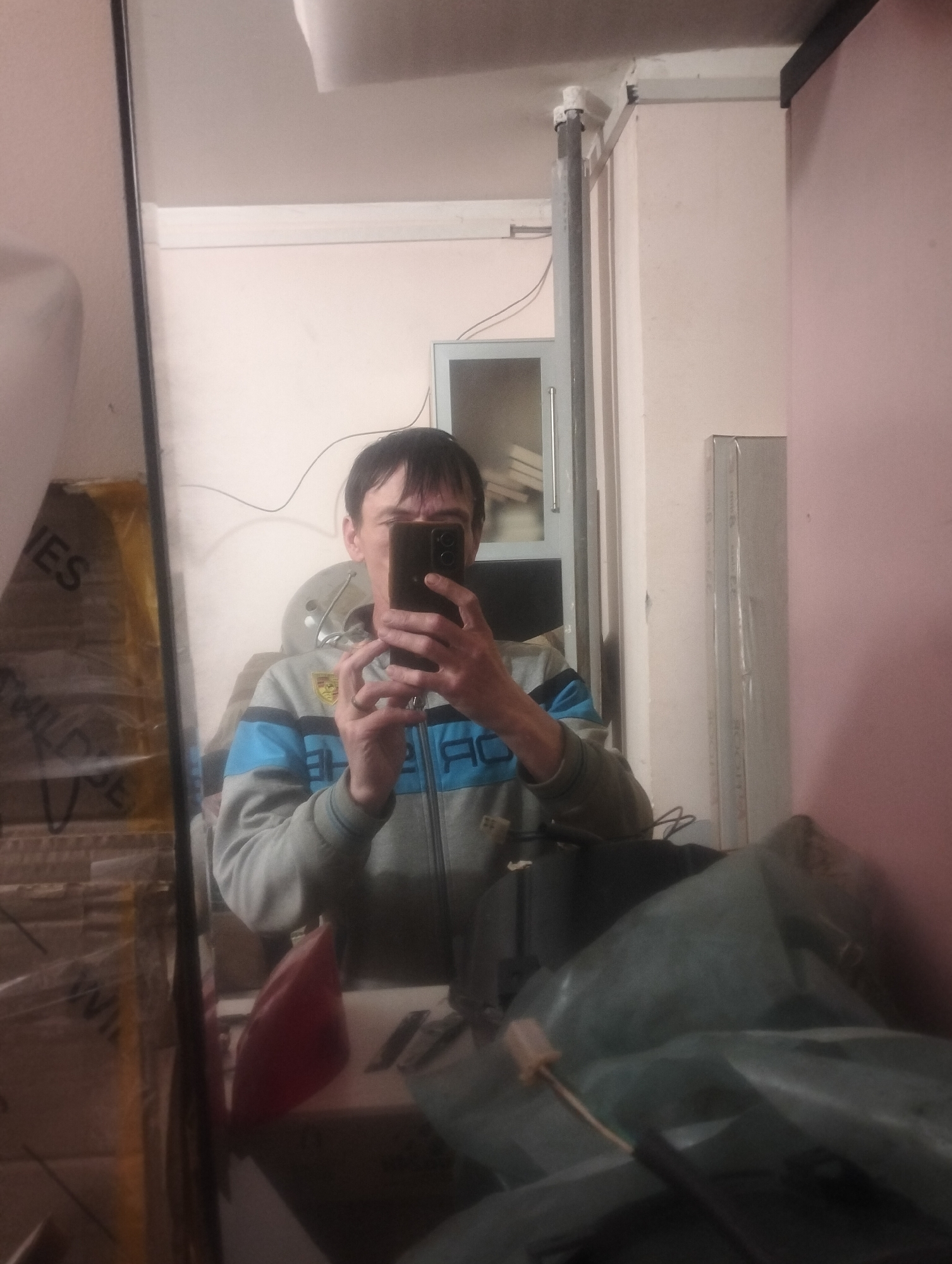 A man is taking a selfie in a mirror in a room. He is wearing a gray zip-up jacket with blue stripes. He is holding a black smartphone in his right hand. In the reflection, behind the man, there are cardboard boxes wrapped in plastic, a red object, and a tall cabinet. The walls of the room are light pink. The floor appears to be covered by a dark green tarp or plastic sheeting. The overall environment suggests a storage area or a workshop. There are no visible indicators to pinpoint the location within the city of Zuyzino, Russia.