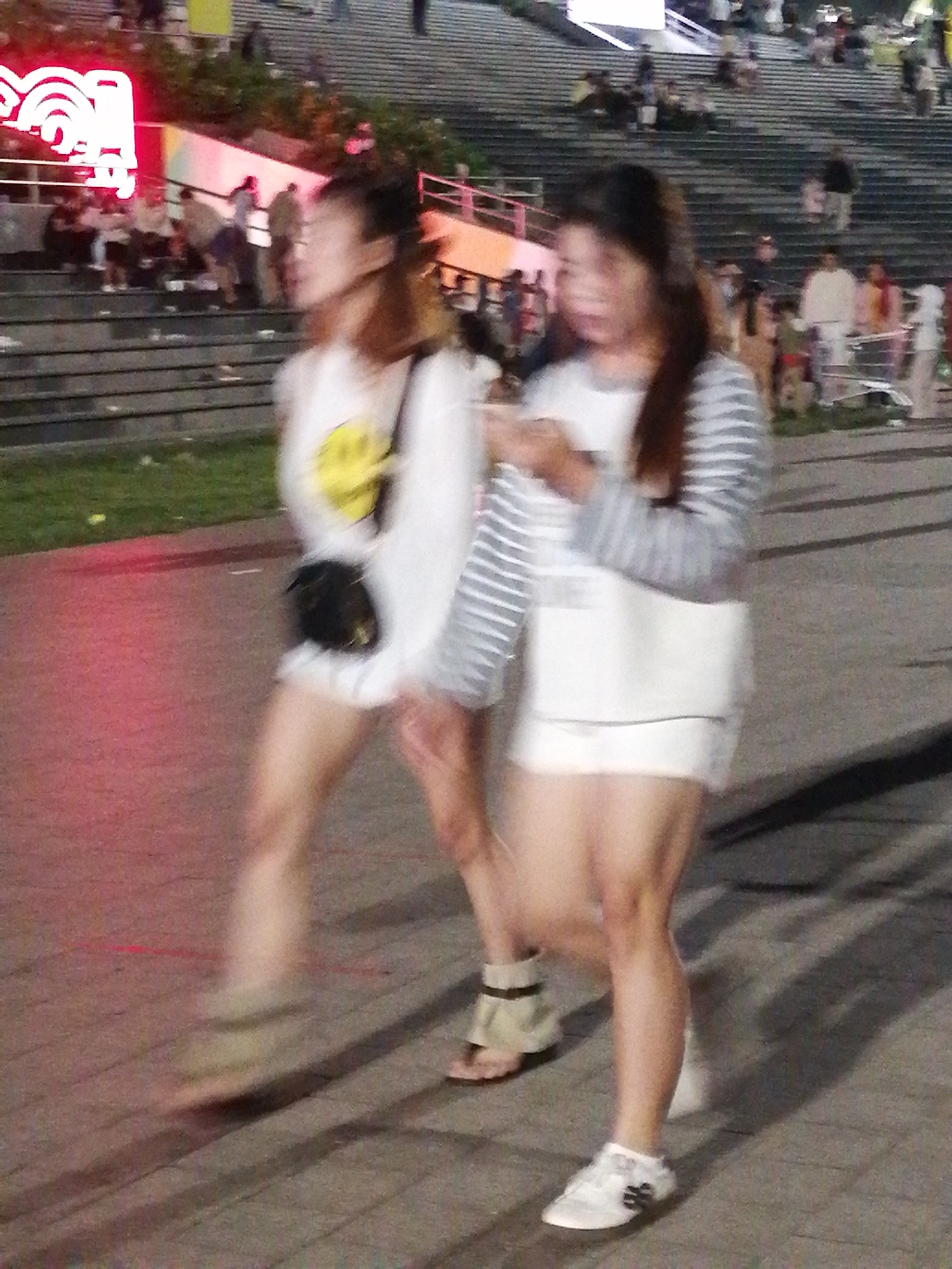 In Da Lat, Vietnam, two young women are walking side-by-side on a paved outdoor pathway. The woman on the left wears a white t-shirt with a yellow graphic and a black crossbody bag, and the woman on the right wears a white t-shirt and a white skirt with a striped long-sleeved shirt over it. They are blurred due to motion, suggesting they are moving at a moderate pace. The environment includes a grassy area to the left and a large set of concrete stairs in the background, on which other people are visible sitting and standing. Neon lights are visible in the upper left corner of the image. The lighting suggests it is evening or night.