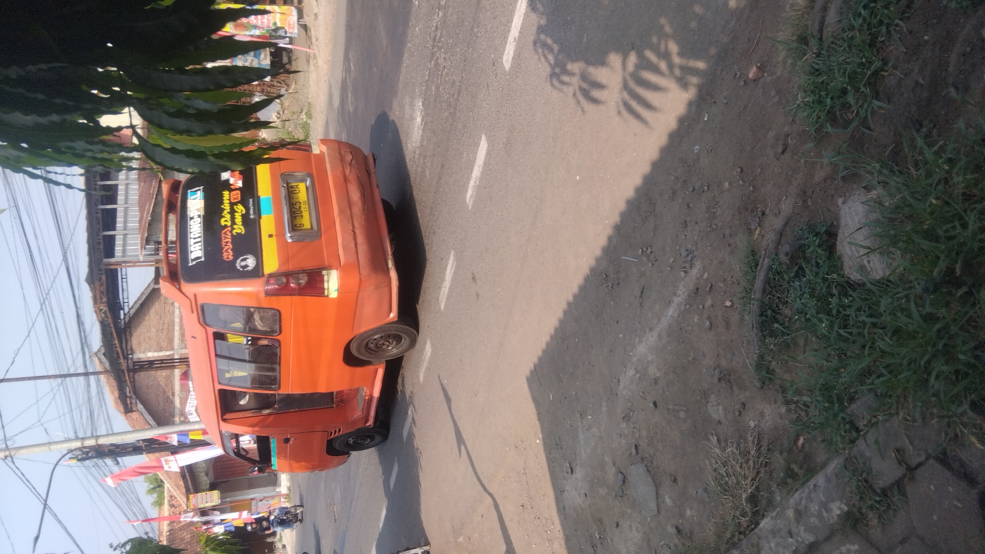 The image depicts an orange public transport van on a street in Proyonanggan Tengah, Indonesia. The van has the text "BATANG-PEKAL" and "HANYA Dirimu Yang Ku" on its rear. A license plate reading "G 1045 OA" is visible. The van is in motion, indicated by the blurred motion of the wheels. The street appears to be paved with asphalt and has white lane markings. To the left of the van, a person on a motorcycle is visible. The background shows buildings with corrugated metal roofs and utility poles with numerous wires. Lush green foliage from a tree partially obscures the right side of the image. The ground beside the road is unpaved and covered with dirt, rocks, and sparse vegetation. The lighting suggests it is daytime with bright sunlight, casting shadows on the road.