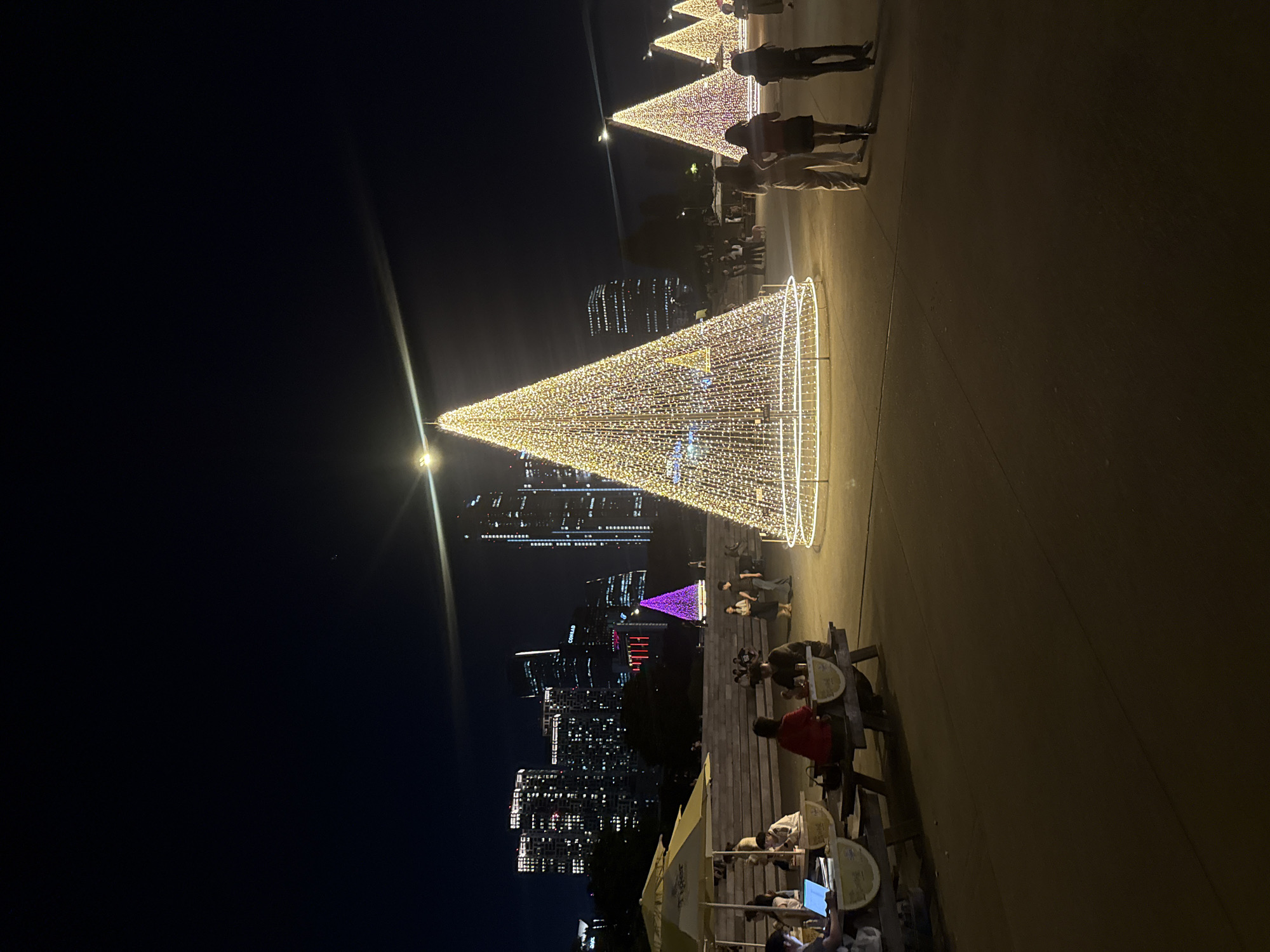 This image shows an outdoor night scene in what appears to be a park or overlook in Seoul, South Korea. Several large, illuminated Christmas tree structures are visible. The foreground features picnic tables with people seated at them. The background displays the city skyline illuminated with lights. The largest Christmas tree is in the center of the image and is covered in yellow lights. A purple Christmas tree is also visible on the left, in front of the city skyline. Some people are standing and observing the scene.