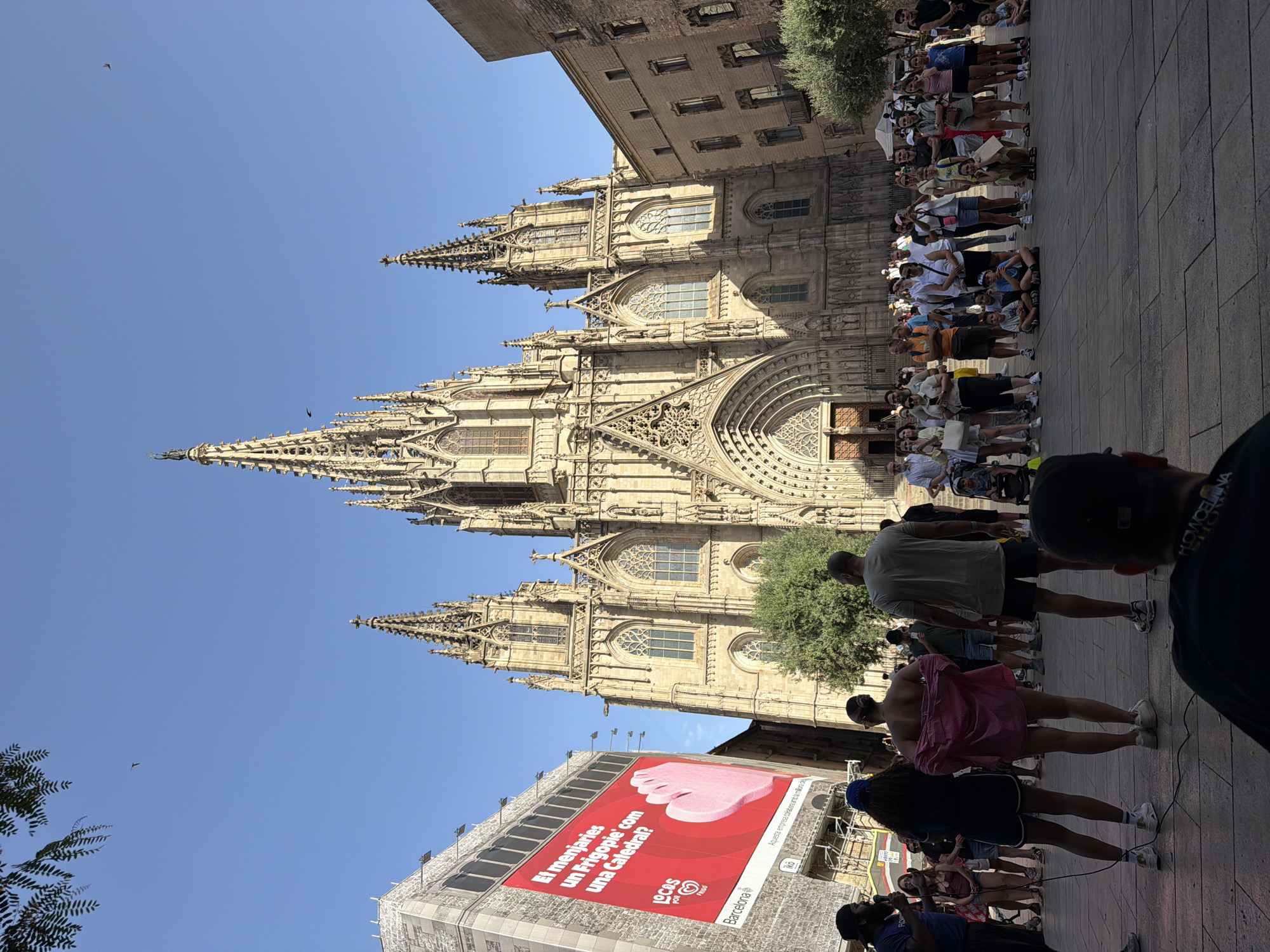 This media file depicts a lively outdoor scene in Barcelona, Spain, dominated by the impressive Gothic architecture of a cathedral. The image is vertically oriented. A large group of people, appearing to be tourists or locals, are gathered in a plaza in front of the cathedral. They are dressed in casual summer clothing, suggesting a warm, sunny day. Some individuals are looking at the cathedral, while others are engaged in conversation or taking photos.
In the foreground, a large, vibrant red billboard is prominently displayed on a building. The billboard features an advertisement with text in Catalan. Visible text includes "El menjaries un Frigopie com una Catedral?", "LOCOS por FRIGO", and "Barcelona". The advertisement also shows a graphic of a pink, ice-cream-like object.
The architecture of the cathedral is intricate, with numerous spires, arched windows, and detailed stonework, indicating it's a significant historical and religious landmark. The clear blue sky above suggests fair weather. The overall atmosphere seems relaxed and communal, with people enjoying the sightseeing and the atmosphere of the city. The presence of the billboard and the crowd suggests this is a public area experiencing a fair amount of activity.