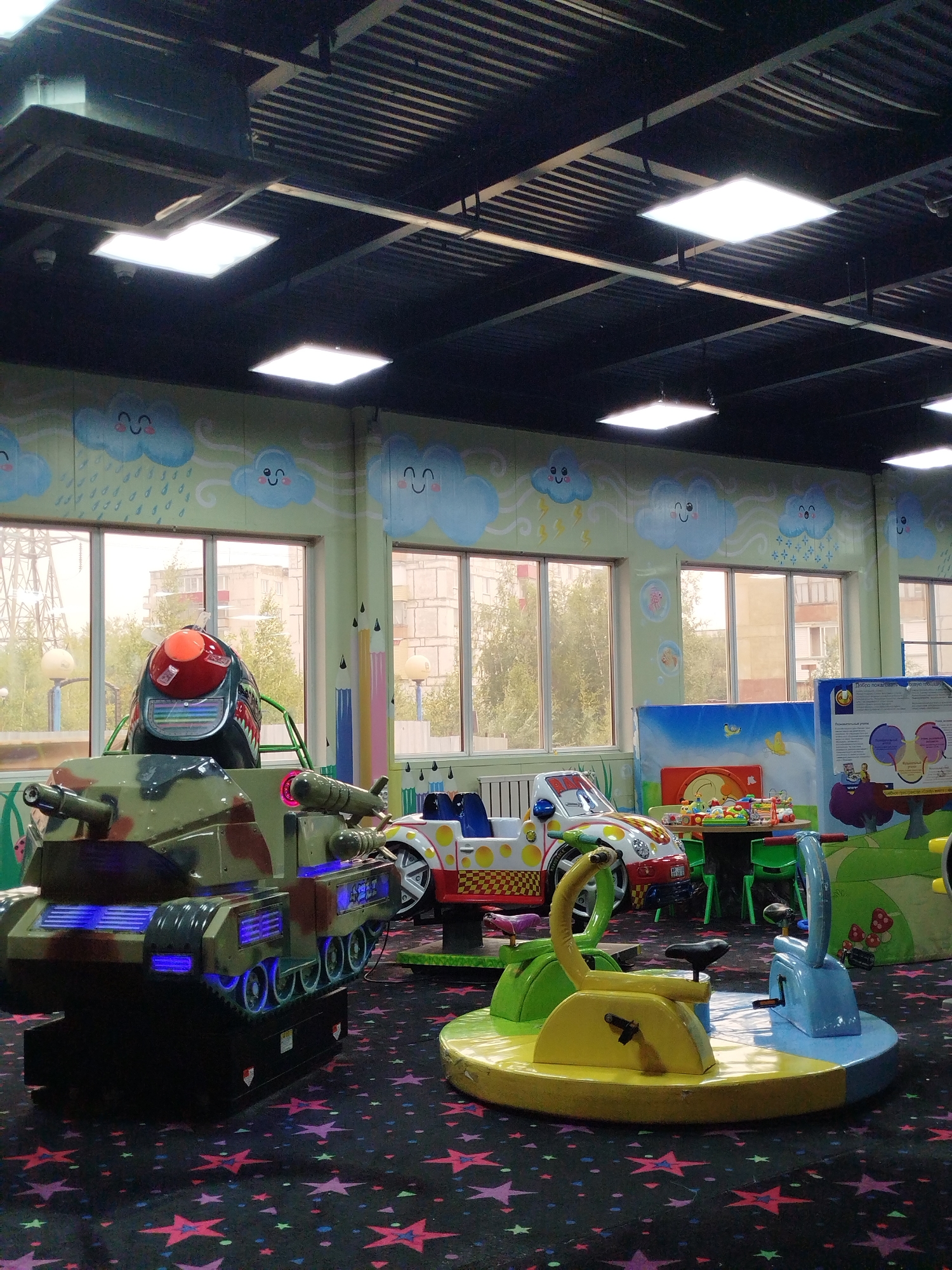 An indoor children's entertainment center is depicted in Mirny, Russia, featuring a brightly colored, cartoon-themed environment. The walls are painted with cheerful clouds and rain, while the floor is covered in a dark carpet patterned with red, pink, and purple stars. In the foreground, a child-sized ride resembling a tank, painted in camouflage with blue illuminated accents, is positioned prominently. To the right, a spinning carousel-like ride with two seats, one yellow and one blue, is visible. Behind these rides, a child-sized car-shaped ride with a yellow and white checkered design and a red stripe is parked. Further back, a circular table with various toys is set up, surrounded by green chairs. Large windows look out onto a street scene with trees and apartment buildings. The ceiling features a dark paneling with rows of fluorescent lighting fixtures. There are no people visible in the image.