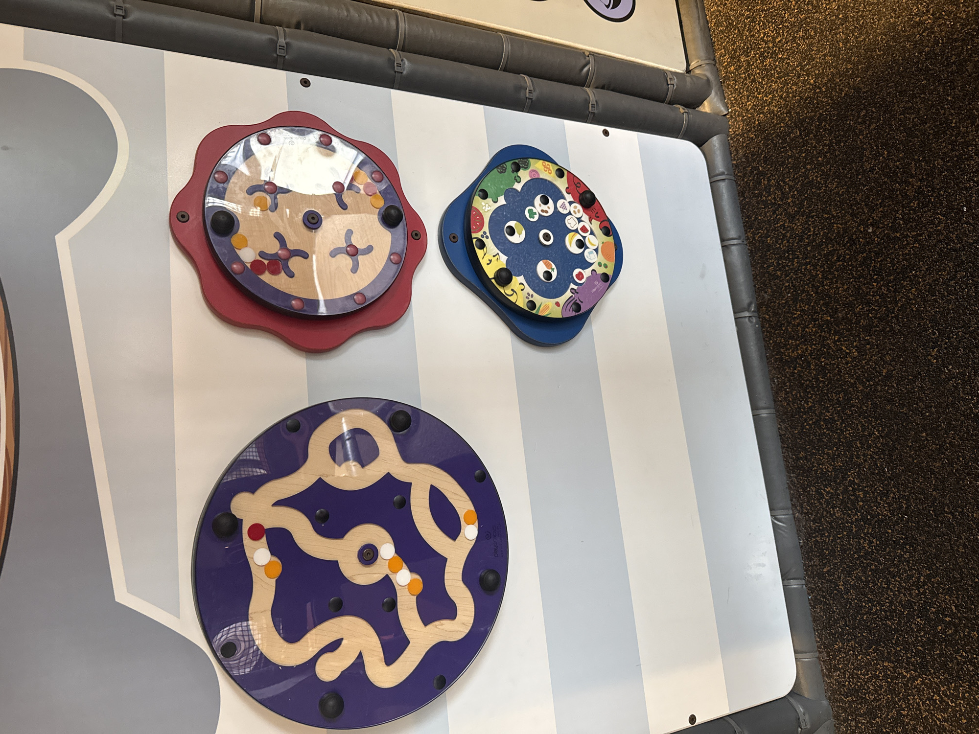 The image shows a play area with interactive wall games. There are three circular games mounted on a white and blue striped wall. The top left game is red and has a clear plastic covering small wooden shapes. The top right game is blue and has a cartoonish food theme. The bottom game is purple and has a wooden track with small balls. The play area is located in Mountain Park, United States of America.