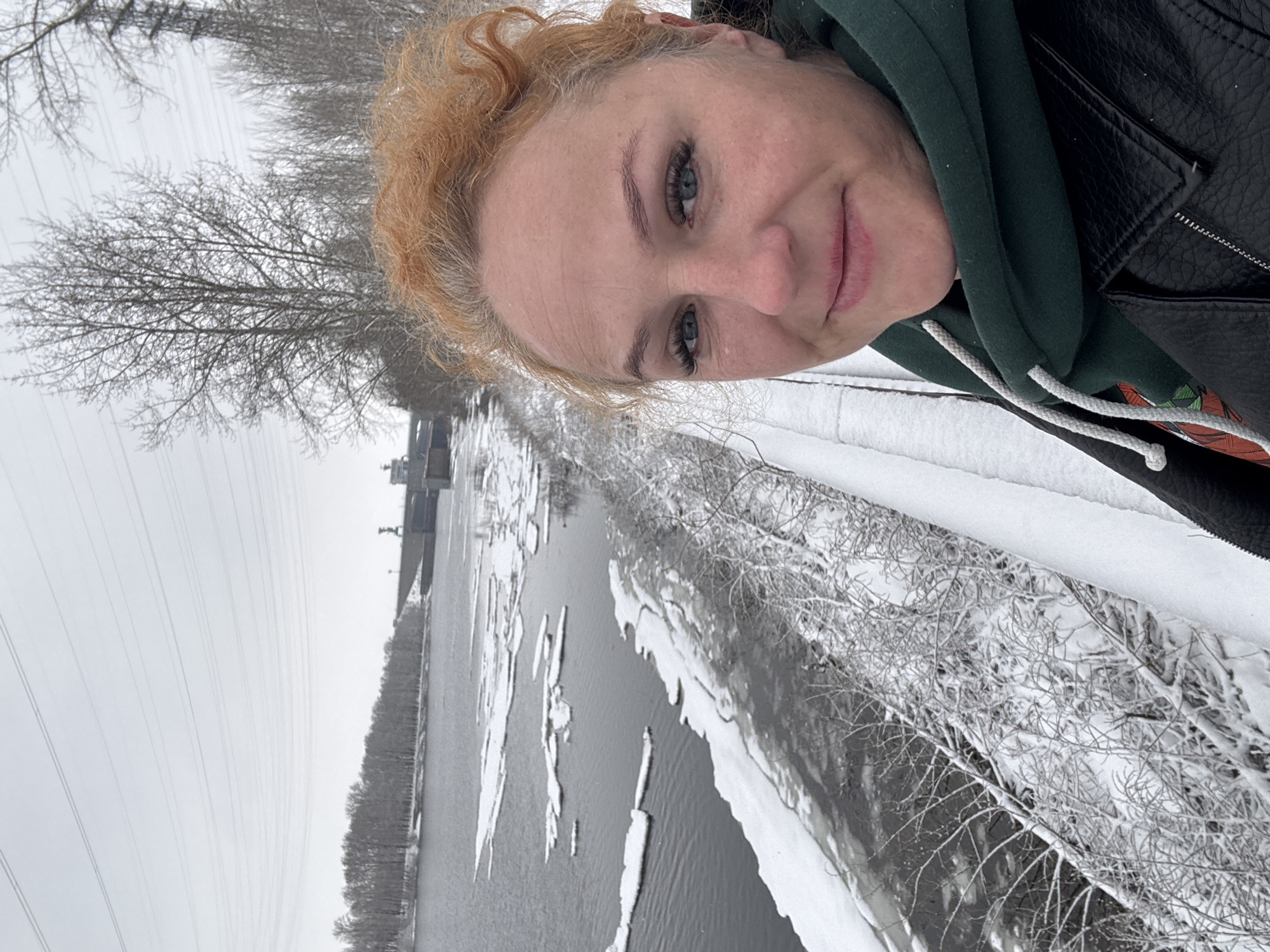 A woman with red hair and light eyes is taking a selfie, in a landscape with snow, frozen river and trees. She is wearing a black jacket with a green hoodie. The river has pieces of ice floating on it. On the other side of the river, there are trees and some type of building. The city is likely Chaikovsky, Russia.