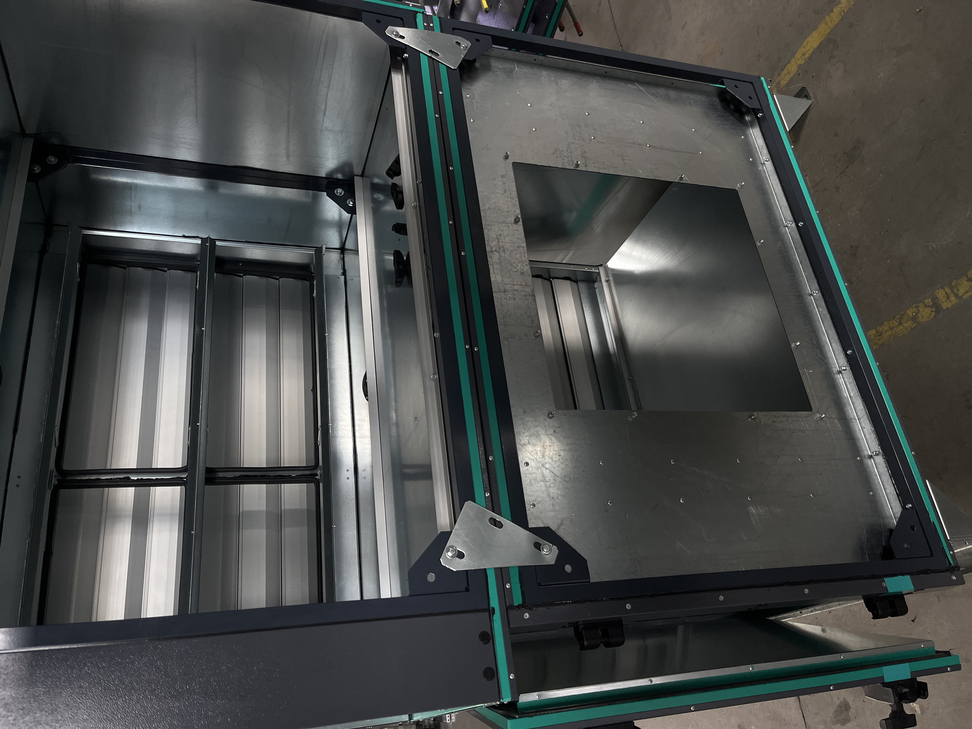 This is a full-shot, eye-level, indoor photograph of an open, industrial metal cabinet or enclosure. The cabinet is constructed of dark grey and galvanized metal. The interior features shelves made of horizontally oriented, reflective metal slats. The exterior of the cabinet is dark grey with green sealant strips along the edges of the open doors and frame. There is a large, rectangular opening in the front lower section of the cabinet. The cabinet is situated on a concrete floor marked with yellow lines, consistent with an industrial or workshop environment. There are no people visible in the image. The lighting is bright and even, suggesting artificial lighting typical of a commercial space. The location is not specified within the city of Kyiv, Ukraine, and no information is available to infer such a location.
