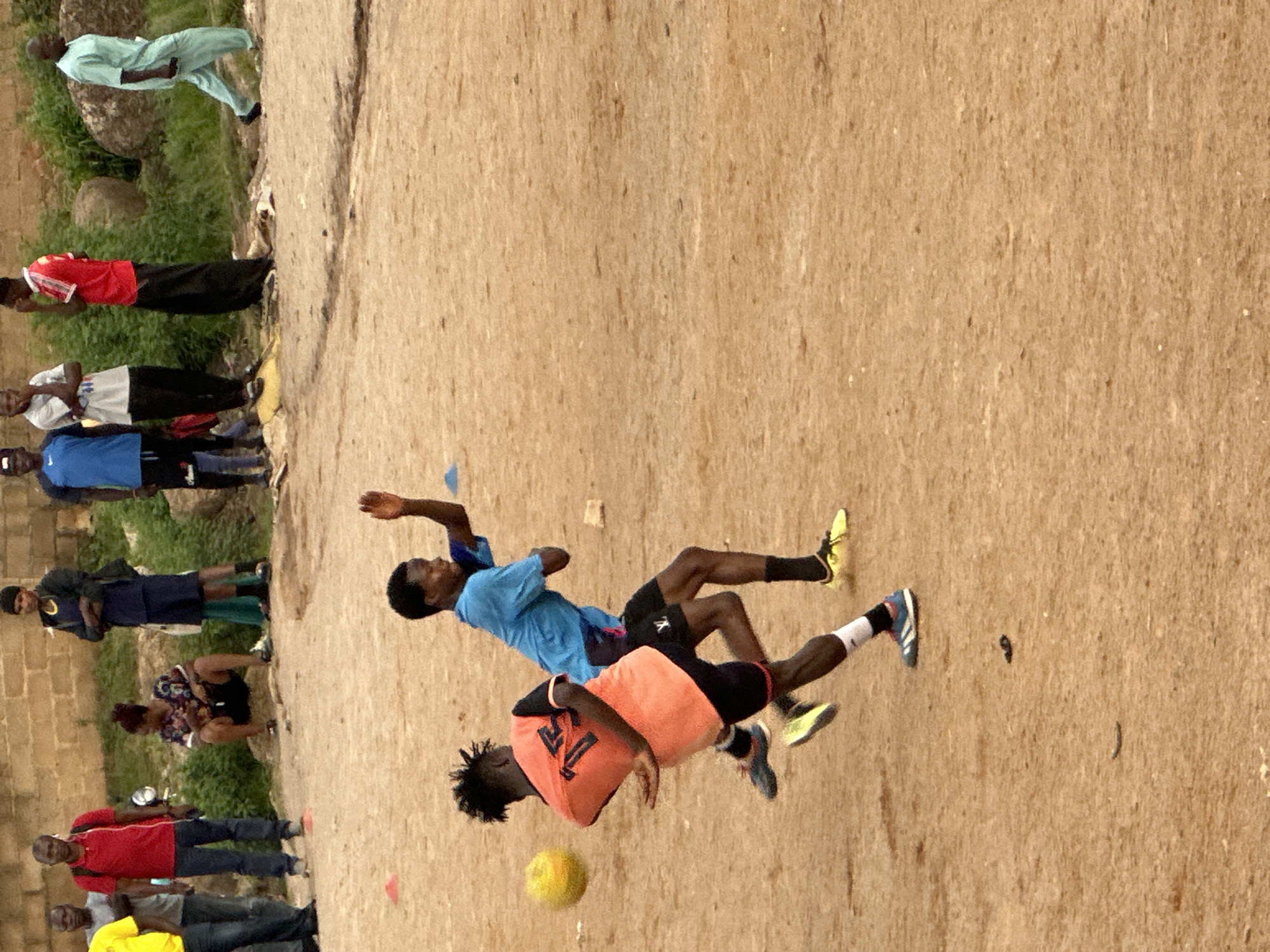The image shows a group of people on a dirt field, likely playing soccer. Two young men are in the foreground, engaged in a play. One player in an orange jersey with the number 77 is being tackled by a player in a blue shirt. A yellow soccer ball is visible on the ground near the orange-jerseyed player.
Several other people are standing and observing the game around the edges of the field. On the left side of the image, a line of individuals are watching. Some are wearing casual clothing like t-shirts and shorts, while others are in athletic wear. One person is wearing a light blue traditional outfit. The background consists of a brick wall and some greenery, suggesting an outdoor setting in a developed area, possibly in Jos, Nigeria, as indicated by the location context. The ground is dusty and uneven. The lighting suggests it is daytime, with natural light illuminating the scene. There is no clear indication of the weather, but the light suggests it's not overcast. The overall atmosphere appears to be one of casual sports activity and community gathering.