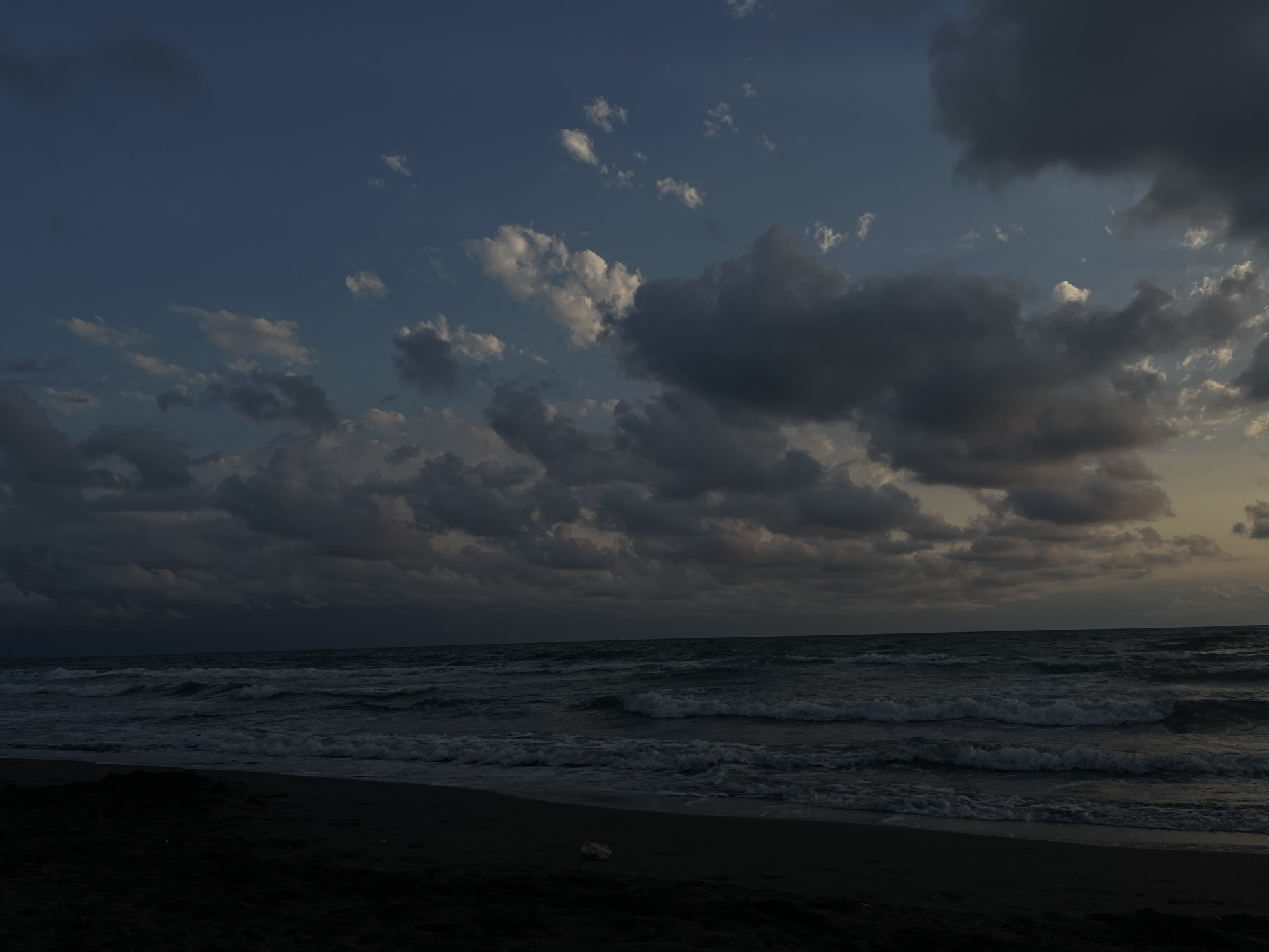 The image depicts a seascape with a dark sandy beach in the foreground, leading to the turbulent waters of the Black Sea under a cloudy sky. The sky transitions from a deep blue at the top to lighter hues near the horizon, with a mix of dark, dramatic clouds and scattered lighter ones. The sea is characterized by choppy waves with whitecaps, suggesting a breezy or stormy atmosphere. The overall mood is somber and somewhat dramatic, indicative of either twilight or overcast weather conditions. No people or specific structures are visible within the frame, emphasizing the natural landscape. This scene could be located along the coast of სუფსის თემი, Georgia, which is known for its Black Sea shoreline.