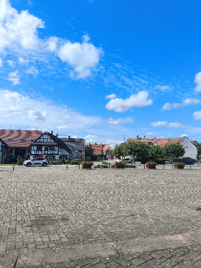 The image depicts a paved square in the village of Kilstett, France, under a clear blue sky with scattered white cumulus clouds. The foreground consists of a grey cobblestone or paver surface. In the midground, a row of approximately eight terracotta-colored rectangular planters with reddish-brown flowers lines an unseen boundary, behind which lies a narrow asphalt street. A white SUV with black trim is parked on this street to the left of the image.
Beyond the street, a collection of traditional Alsatian-style buildings is visible. The prominent structures to the left include multi-story houses with dark brown half-timbering on white facades and reddish-brown tiled roofs. Some windows feature dark shutters. Further right, more residential buildings with tiled roofs and varying architectural styles extend into the background. Green foliage from trees is visible interspersed among the buildings, particularly around the center-right. A black street lamp post stands near the center of the frame, and a red fire hydrant is visible to its right, behind the planters. No individuals are present.