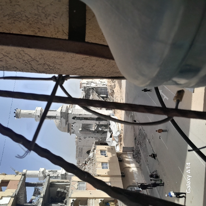 This image depicts a street scene in Khan Yunis, Palestinian Territory, characterized by widespread structural damage. The central focus is the Grand Mosque of Khan Yunis, identifiable by its two minarets and central dome, which exhibits severe damage including large holes in its walls, collapsed sections, and exposed interior structures. Surrounding the mosque are numerous multi-story buildings, all showing extensive destruction with collapsed floors, exposed rebar, and large amounts of rubble.
The paved street in the midground contains scattered debris. On the left side of the street, four adult males are present; some are standing, others are squatting near a bicycle. Three children are visible on the street; one walks towards the foreground, another stands further down the street, and a third is partially obscured by rubble.
In the immediate foreground, a dark, twisted metal grate or window bars partially frame the view. A large, dusty, light blue plastic water cooler bottle is partially visible on the right side of the foreground. The sky is clear.