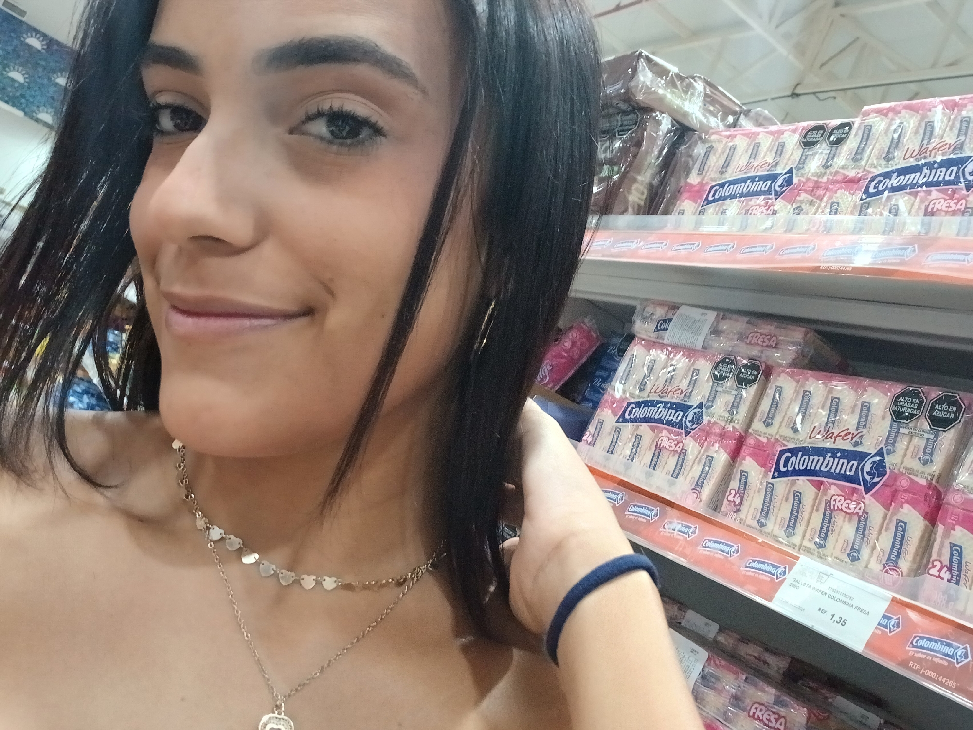 A woman with dark hair and a multi-strand heart necklace is taking a selfie in a store aisle in Juan Griego, Venezuela. The shelves behind her are stocked with "Colombina" wafer cookies. A price tag indicates the wafers cost 1.35 (currency not specified). She has a dark blue elastic band on her wrist.