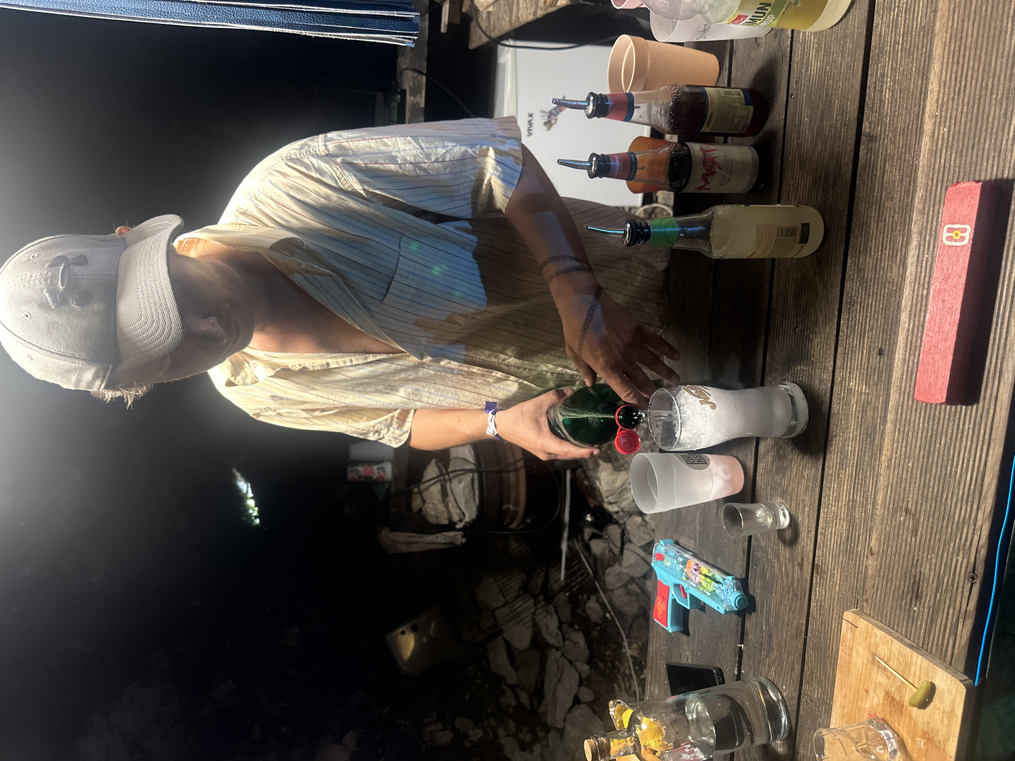 In the city of Bukovik, Montenegro, a person wearing a light-colored, vertically striped button-up shirt and a grey baseball cap is pouring a clear liquid from a green bottle with a red cap into a glass filled with ice. The person has tattoos on their forearm that resemble a chain. They are standing behind a rustic wooden table laden with various items.
On the table, to the left of the pouring action, is a clear bottle with a cork stopper and yellow liquid, a black smartphone, and a blue and red toy water gun. Closer to the glass being filled are a frosted white cup and a small shot glass. To the right of the glass, there are several bottles with pour spouts, including one with a green cap and another with a red cap, and two orange cups. In the background, there is a dark, rocky environment, a wooden barrel, and a white appliance that appears to be a small refrigerator. A bright light illuminates the scene from above and to the left, creating strong shadows.