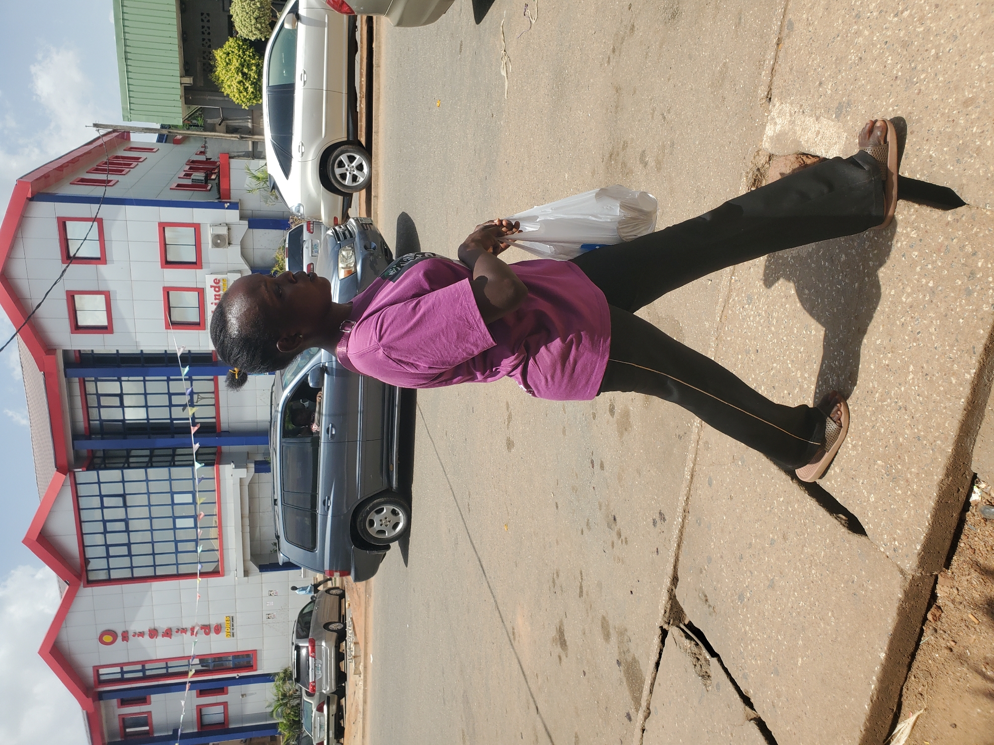 An adult woman stands on a concrete sidewalk, dressed in a short-sleeved purple shirt and black pants with a gold-colored stripe along the side. She faces the camera's left, holding a white plastic bag. The sidewalk is cracked near the curb, and a shadow is cast behind her.
Behind the sidewalk, a street shows several parked cars. The building behind the cars is white with red trim around the roof and windows, with blue accents, and has lettering on its facade, including Chinese characters. The sign reads "Diamond Super Store" or similar. Based on the style of building and the street scene, the image appears to be taken in Jos, Nigeria.