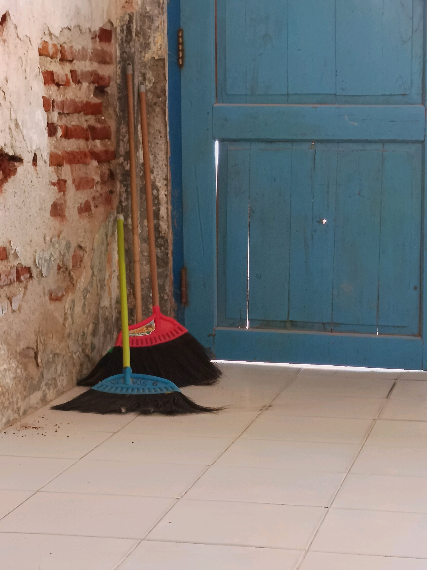 The image depicts an outdoor scene against a weathered brick wall and a blue wooden door. In the foreground, on a tiled floor, are two brooms. One broom has a bright yellow handle and a blue brush head, while the other has a red brush head and a wooden handle. The brooms are positioned near the base of the wall, with the blue-doored structure to the right. The wall shows signs of age, with exposed brick and peeling plaster. The blue door appears to be made of vertical planks and has a visible hinge on the left side. The lighting suggests it is daytime, casting shadows on the floor. The scene appears to be located in Labuhan, Indonesia, given the context provided.
