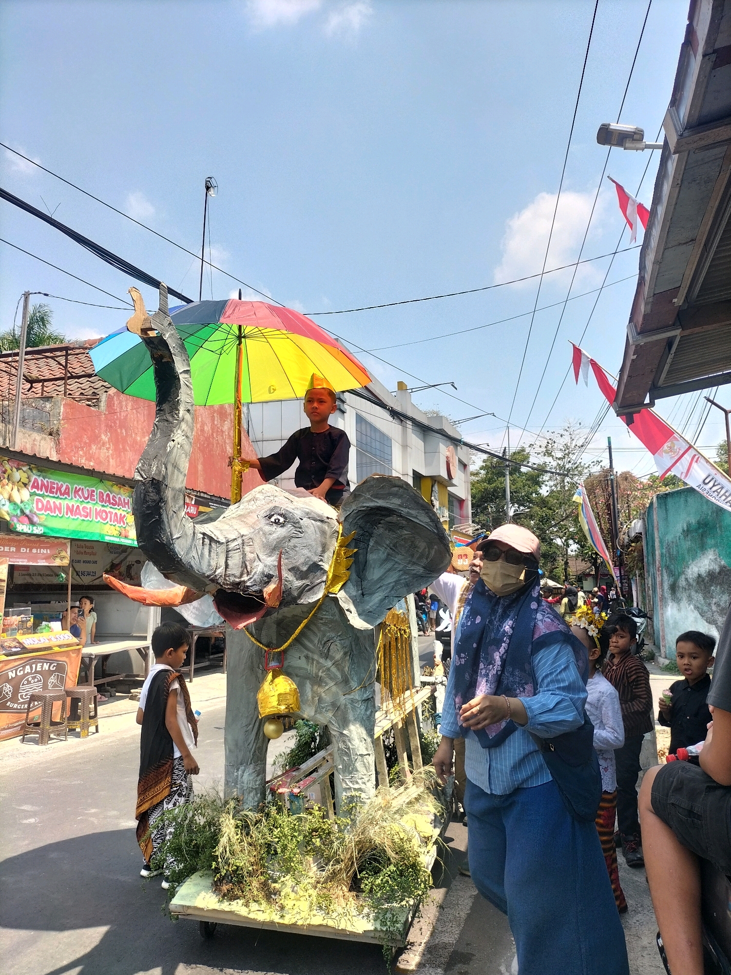 In Malang, Indonesia, a street festival is underway with a large, papier-mâché elephant float as the central focus. A young boy, wearing a dark outfit and a golden cap, sits atop the elephant's head, holding a rainbow-colored umbrella. The float is adorned with gold accents and a large bell, and is decorated with greenery at its base. A woman wearing a blue headscarf and mask, along with a blue striped shirt and wide blue pants, stands beside the float, looking towards the right. Other children are visible in the background, some observing the scene. The street is lined with buildings and shops, and red and white Indonesian flags are displayed, indicating a patriotic celebration. The weather appears to be sunny, with a clear blue sky overhead.