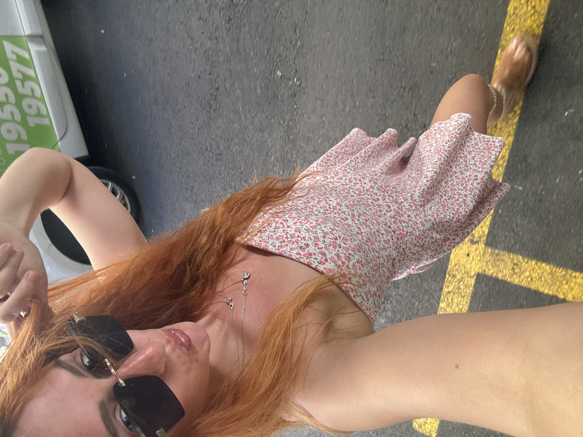 A female individual is depicted in a selfie taken on an asphalt road in Budva, Montenegro. She possesses long, reddish-brown hair and fair skin, and is wearing dark sunglasses with gold-colored accents, two thin necklaces, and a light-colored strapless dress with a pink floral pattern. Her lips are puckered in a pose, and her left hand is raised towards the top of her head. Her right arm is extended, holding the camera. Her right leg is visible, adorned with a light-colored ankle bracelet and light brown, textured open-toed footwear. In the background, a white vehicle with green numerical text "19550 19577" is partially visible. The ground is dark grey asphalt marked with a prominent yellow painted line. The scene is outdoors, under natural light.