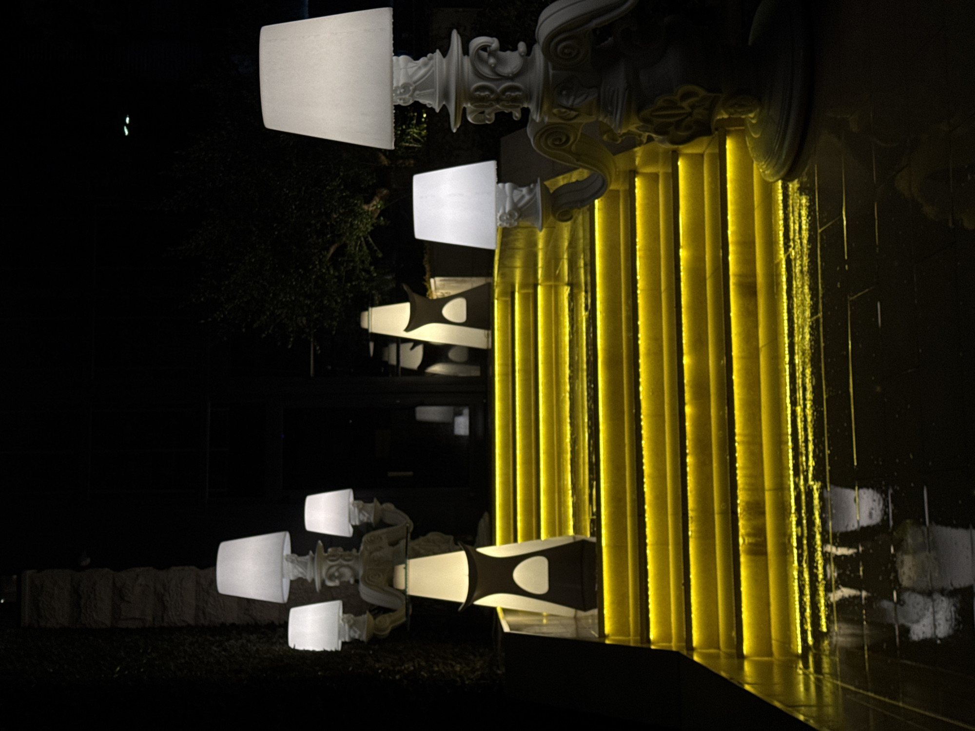 The photograph, taken at night in Budva, Montenegro, features illuminated outdoor decor. In the foreground, two pairs of ornate white lamp posts with square shades flank an object with a white oval accent. The background showcases a wall of vertical golden lights reflecting onto a wet surface. A dark exterior structure and some vegetation are visible.