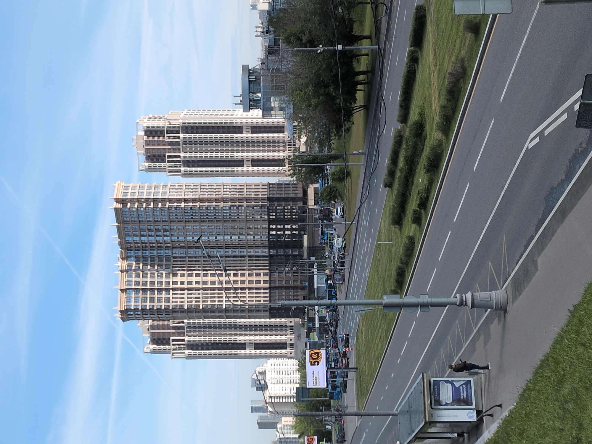The image depicts a street scene in Moscow, Russia. Dominating the left side of the frame are several modern, high-rise apartment buildings with repeating window patterns and layered architectural details. The sky is blue with sparse, faint cloud formations.
To the right of the buildings is a wide, multi-lane road with a median strip of greenery. Cars and buses are visible on the road, moving in both directions. A row of trees lines the road further back.
In the foreground, on the right side, there is a sidewalk with a grassy area. A man is lying on the sidewalk near a bus stop advertisement. Streetlights are also visible along the sidewalk.